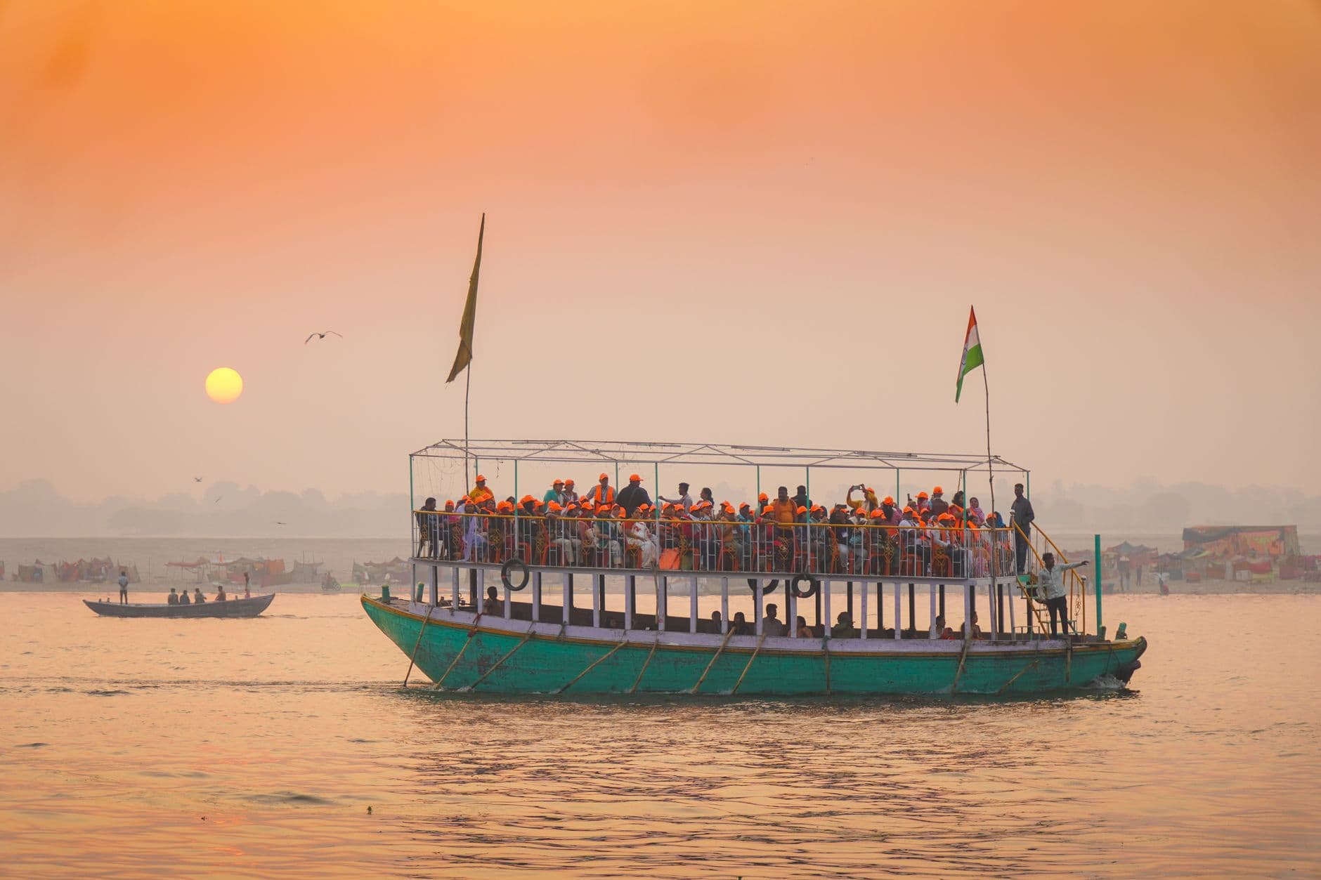A crowded tourist boat filled with orange-clad passengers on the Ganges River at sunrise or dawn, with an Indian flag flying from the vessel, smaller wooden boats nearby, and hazy riverside buildings visible through morning mist. - Photo by Arto Suraj on Pexels