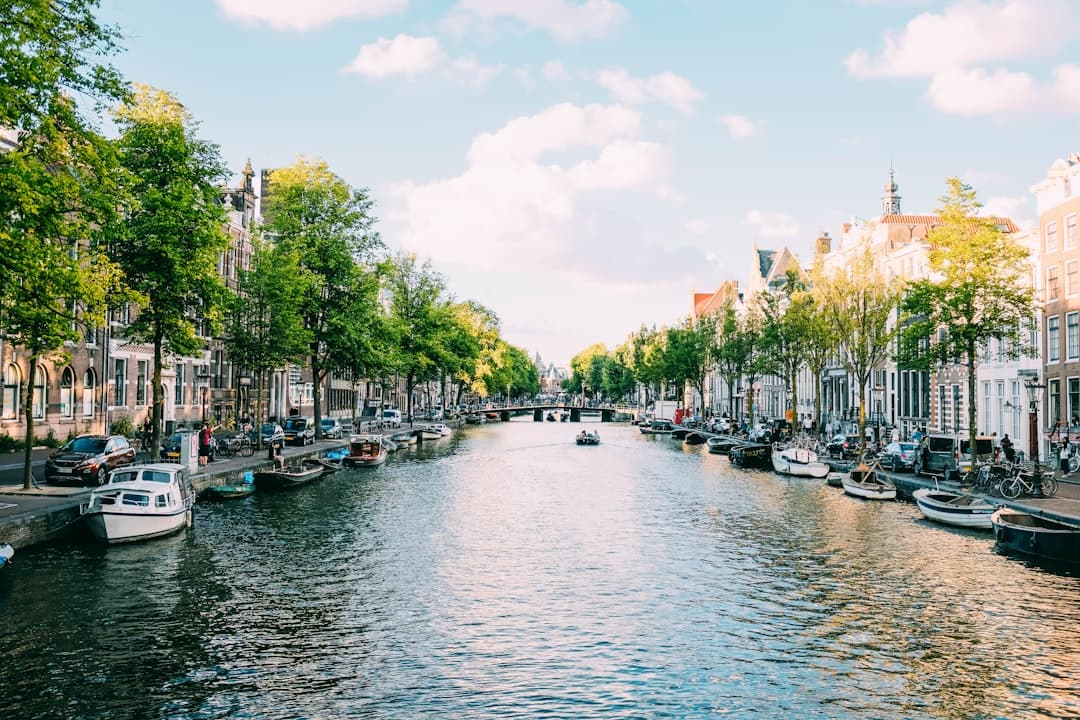 A scenic canal in Amsterdam lined with characteristic Dutch architecture, boats, and tree-lined streets under a partly cloudy sky. - Photo by Adrien Olichon on Unsplash
