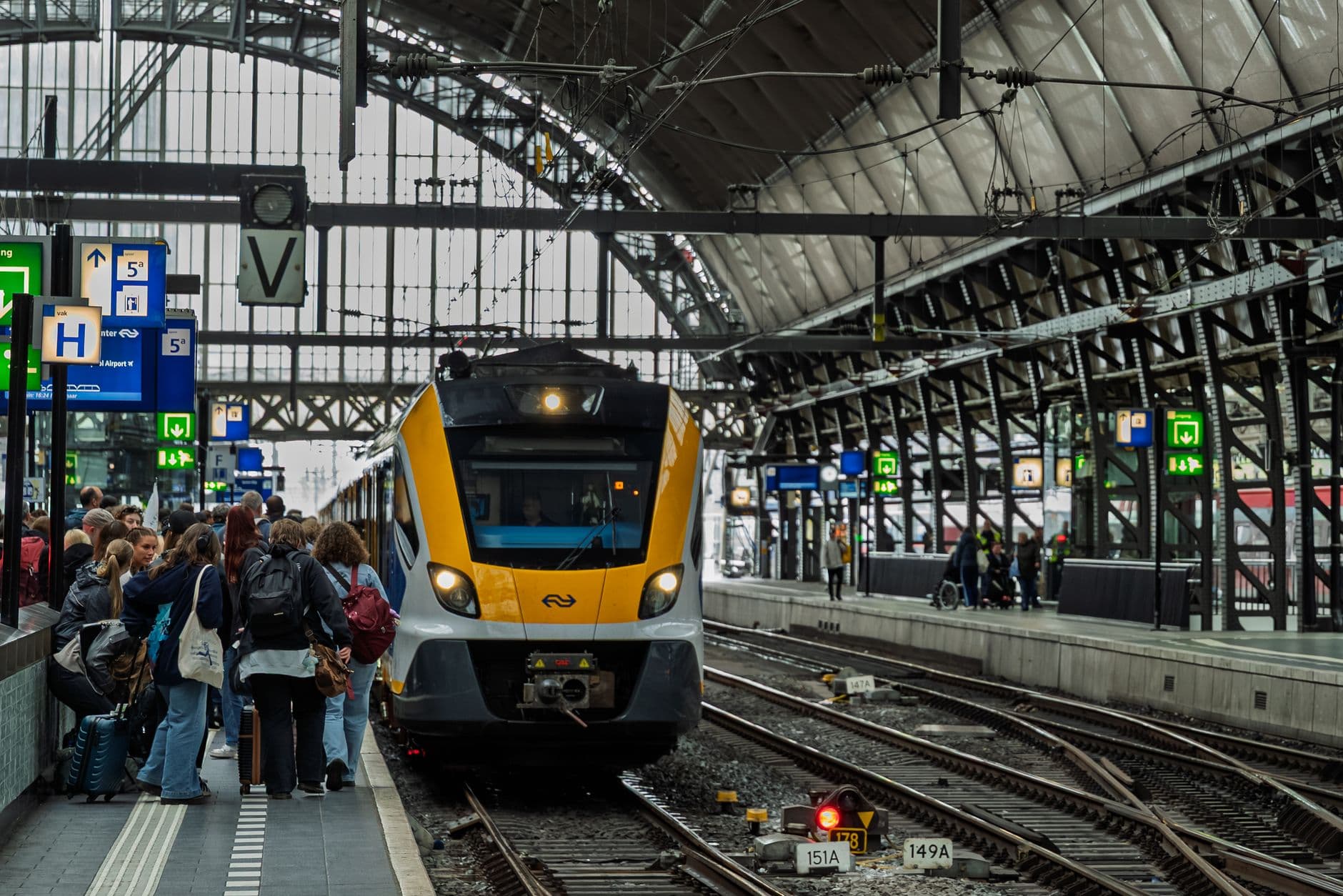 Passengers board a yellow-and-blue NS regional train at a covered railway platform with characteristic Dutch station infrastructure and signage. - Photo by Martijn Stoof on Pexels
