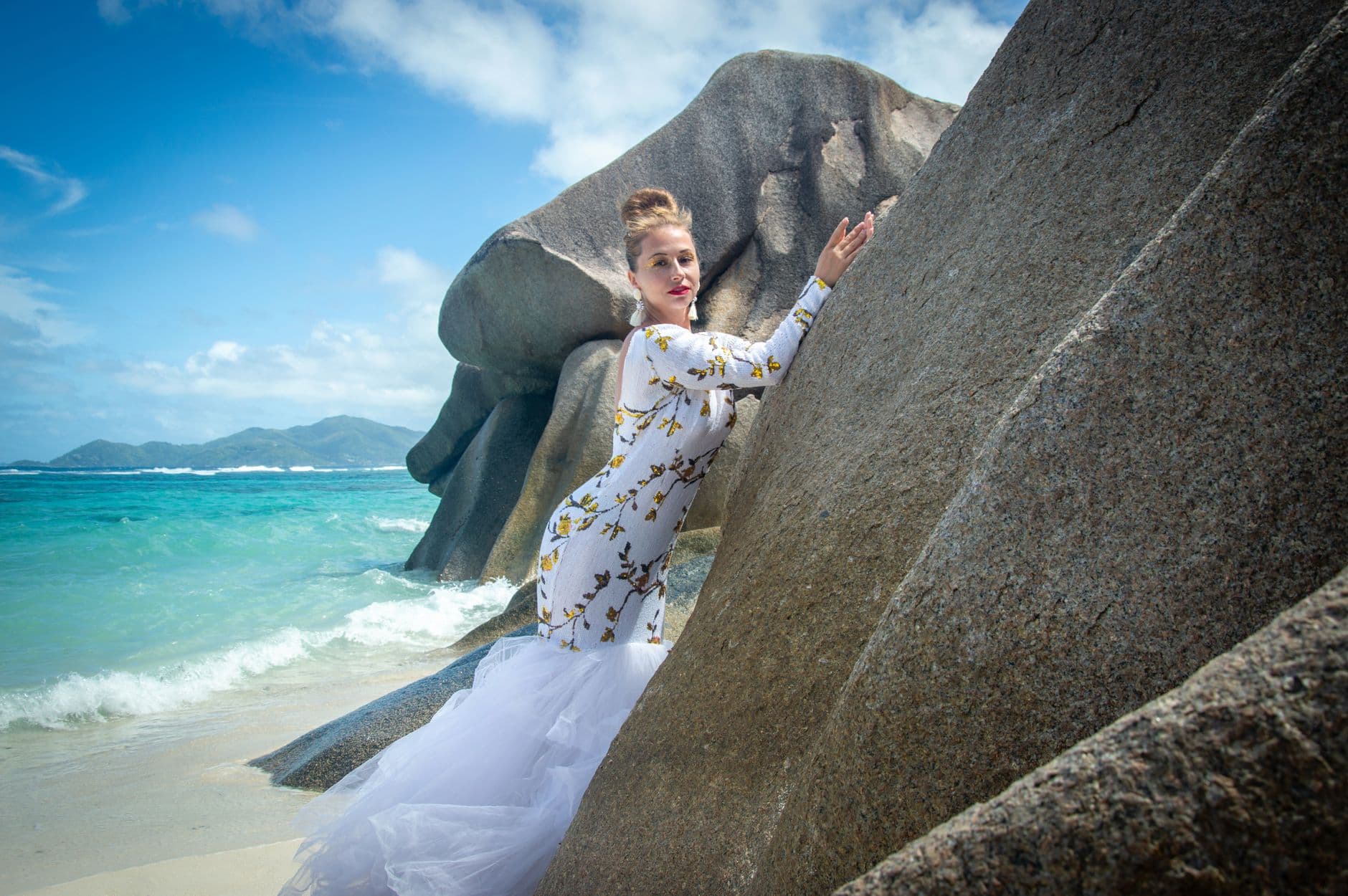 A woman in a white dress with gold floral embroidery poses against massive granite boulders on a pristine tropical beach with turquoise waters and distant islands visible. - Photo by Vika Glitter on Pexels