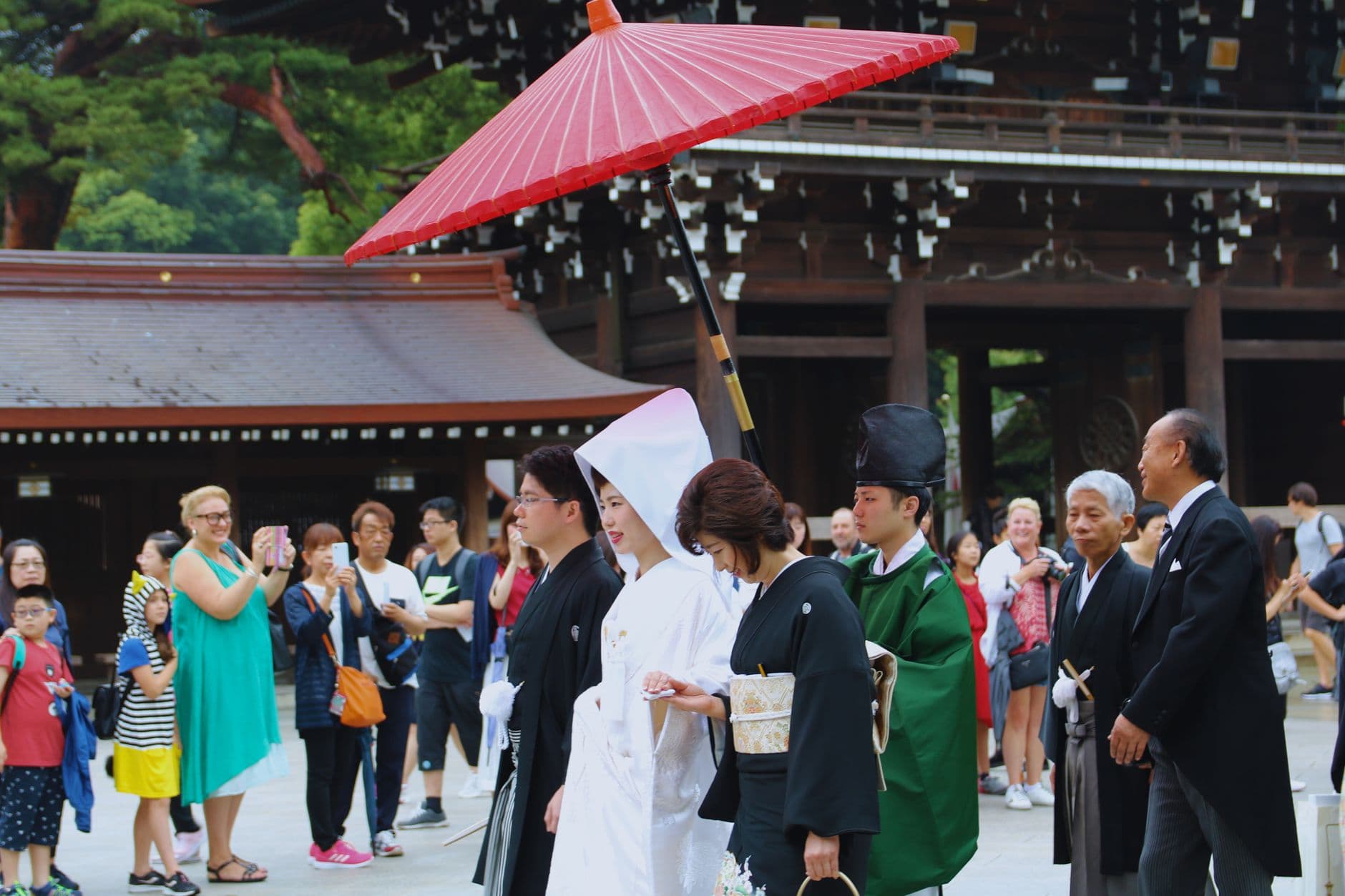 A formal ceremony or event taking place in the courtyard of a traditional Japanese Shinto shrine, with participants in traditional Japanese formal attire (including a bride in white shiromuku wedding kimono and matching white umbrella) and numerous guests and onlookers in the shrine grounds. - Photo by Bruna Santos on Pexels