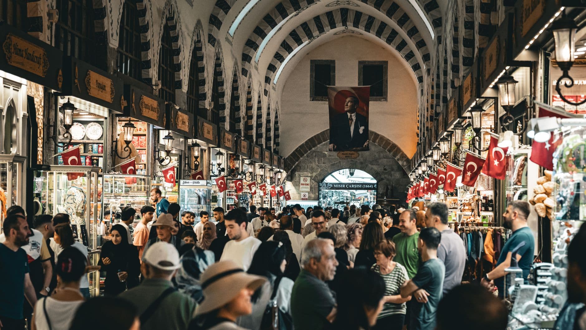 Crowded interior of the Mısır Çarşısı (Egyptian Bazaar) in Istanbul showing the famous vaulted marketplace with Turkish flags, traditional lanterns, vendor stalls, and tourist crowds flowing through the historic arcade. - Photo by Tolga deniz Aran on Pexels