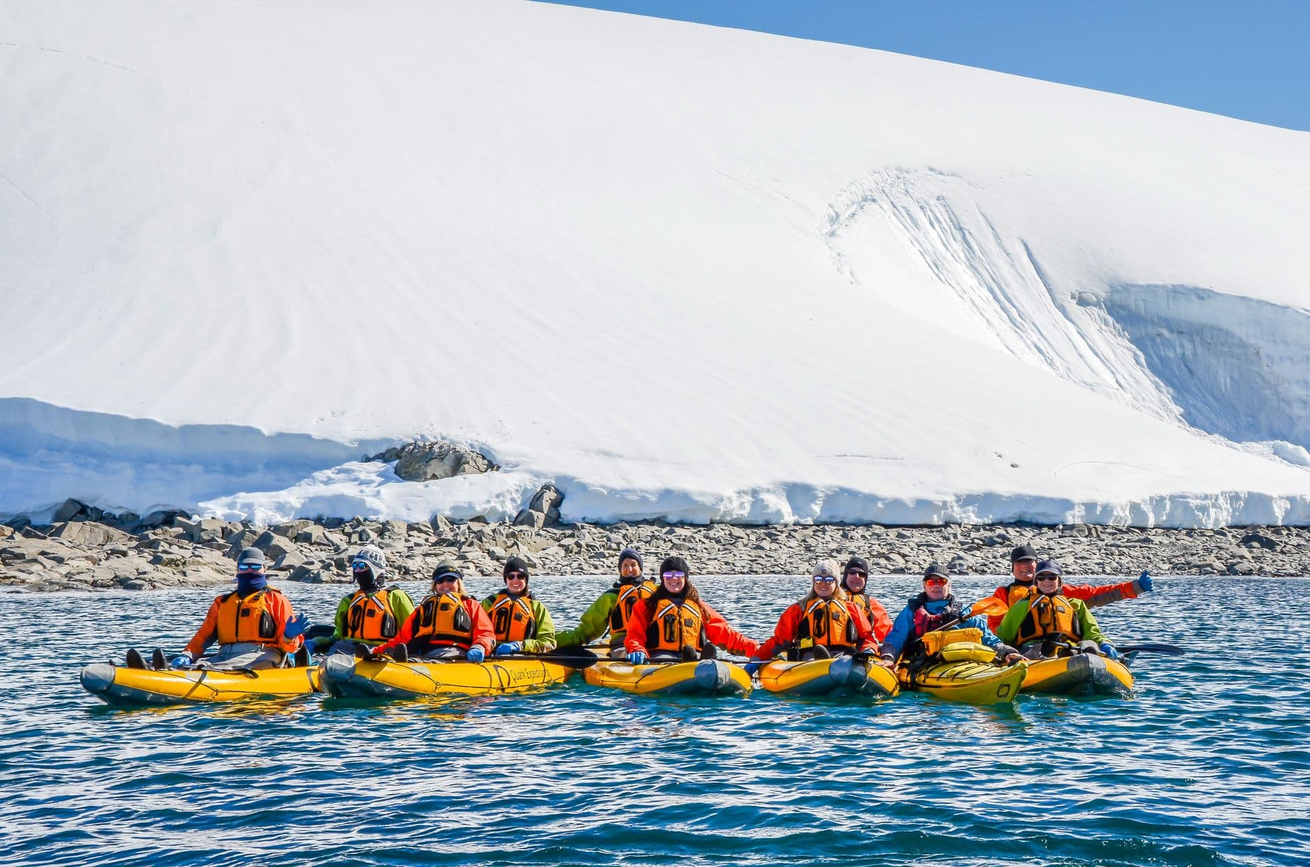 A group of approximately 12 kayakers in yellow inflatable kayaks and orange/red life jackets pose in calm water before towering icebergs and a snow-covered mountainside with visible ice cliffs. - Photo by Karson on Pexels