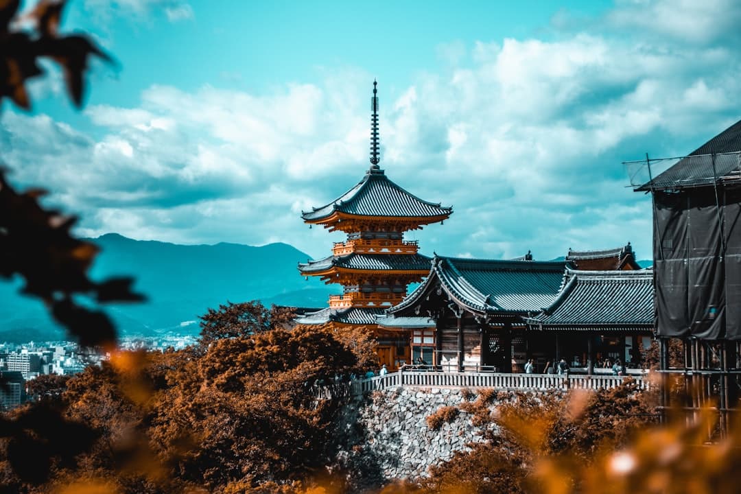A multi-tiered Buddhist pagoda with black-tiled roofs and golden-brown wooden structures dominates the composition, set against mountains and cloudy sky, with autumn foliage in the foreground and urban development visible in the distance below. - Photo by Cosmin Georgian on Unsplash