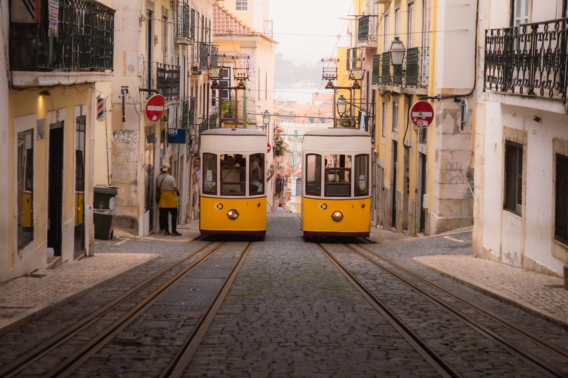 Two yellow vintage trams (Tram 28) ascending a steep cobblestone street in Lisbon's historic Alfama district, with traditional Portuguese buildings on either side and a distant view toward the Tagus River. - Photo by Naimish Verma on Pexels