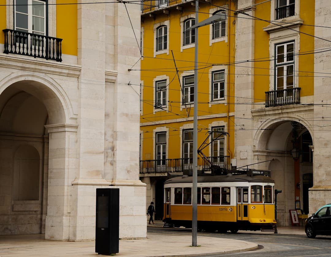 A yellow vintage tram passes through a plaza lined with neoclassical buildings featuring cream-colored stone facades and a striking yellow building in Lisbon's Baixa district. - Photo by Alai Photography on Unsplash