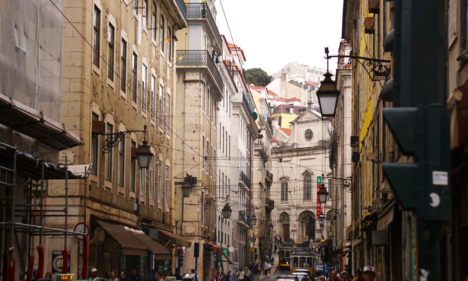 A pedestrian street in central Lisbon lined with historic buildings, featuring a yellow tram, traditional street furniture, and a church visible at street's end. - Photo by Emma on Pexels