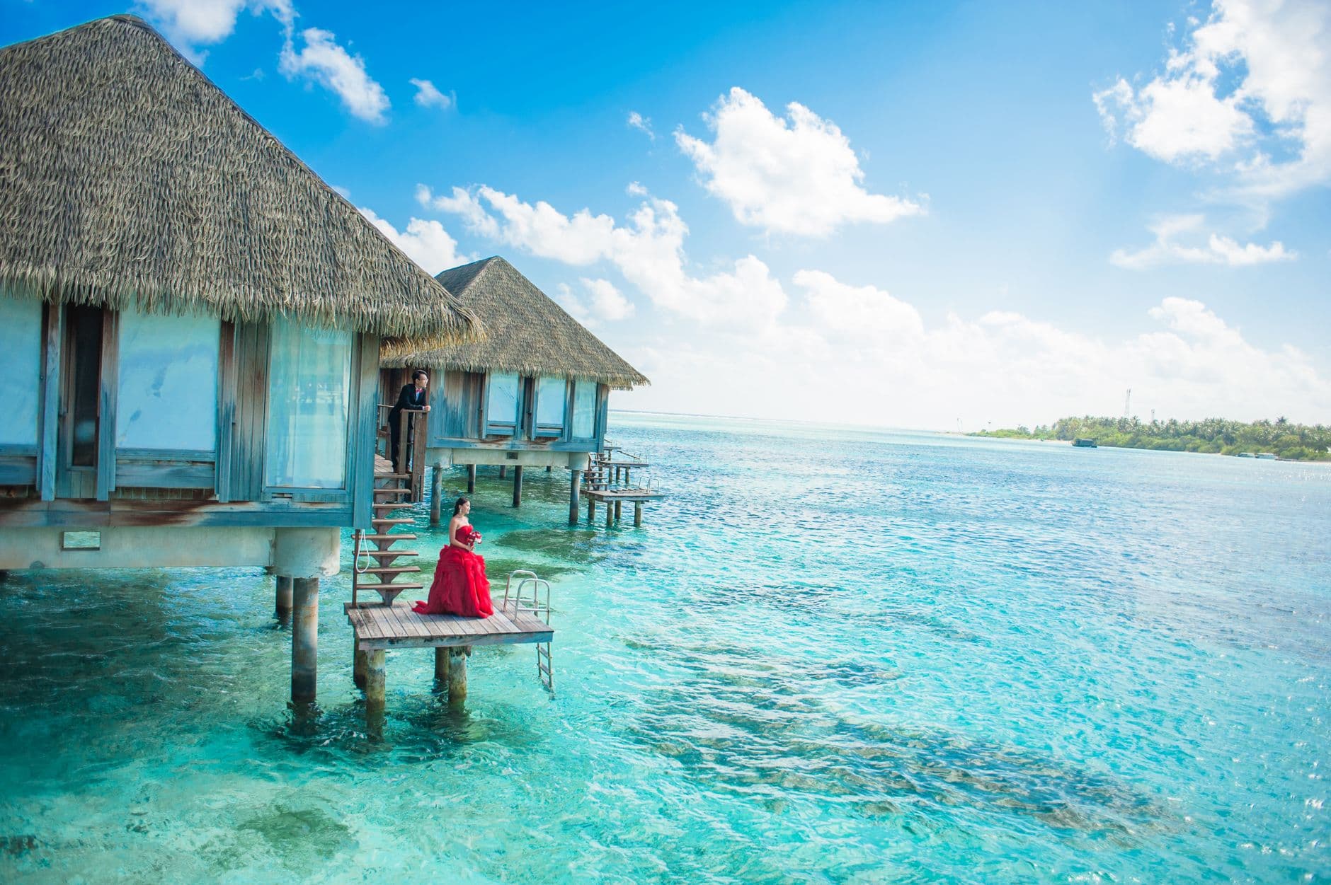 A woman in a red dress sits on a wooden platform extending from turquoise over-water bungalows with thatched roofs in a crystalline lagoon, with staff visible in the resort structures. - Photo by Asad Photo Maldives on Pexels