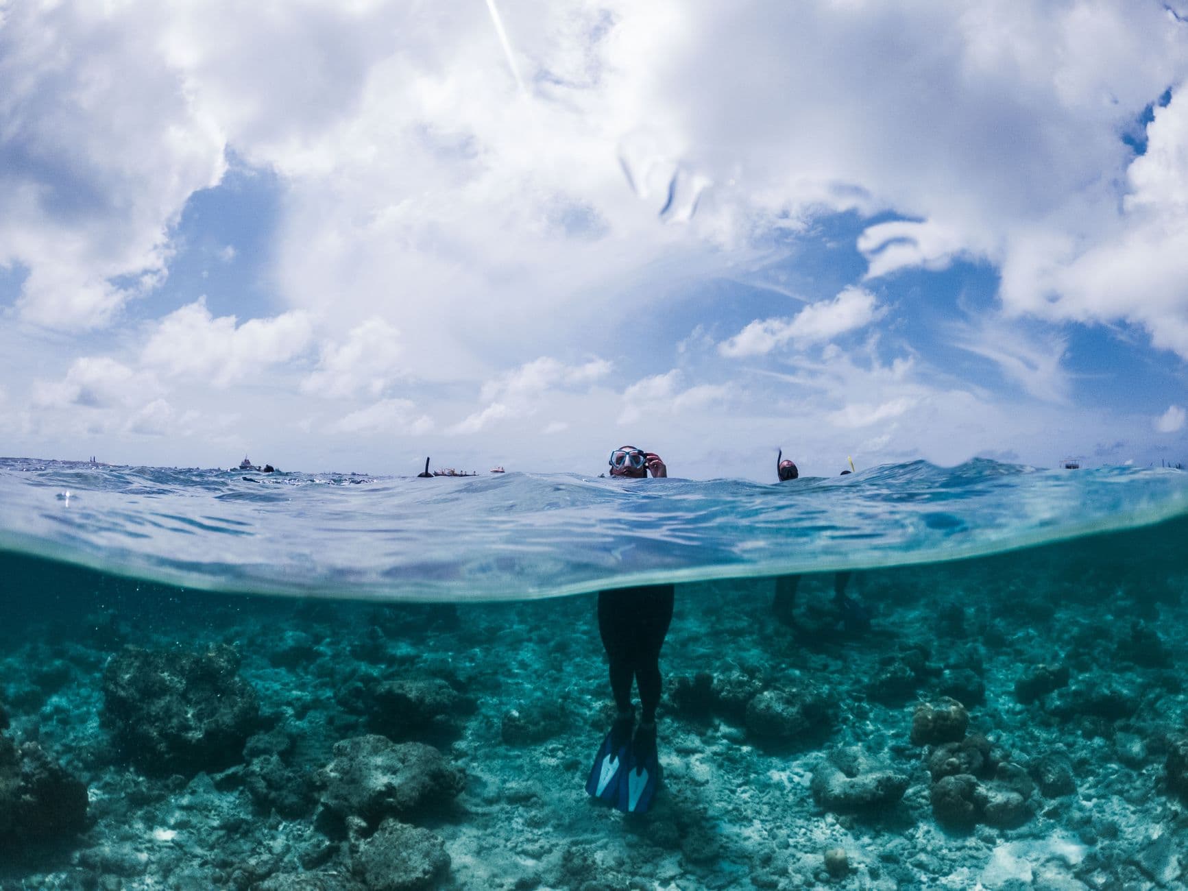 A split-level underwater photograph taken from a snorkeler's perspective showing a diver in the foreground exploring a healthy coral reef with traditional Maldivian dhoni boats visible on the surface above. - Photo by Maahid Photos on Pexels