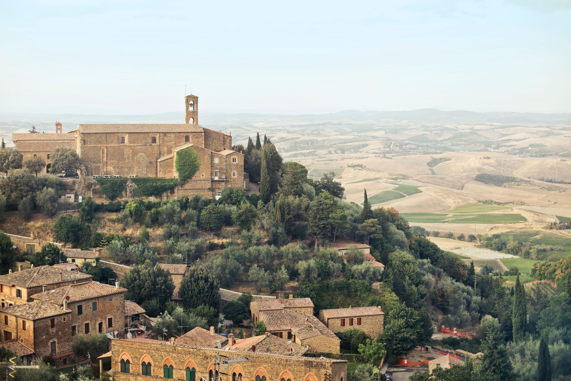 A hilltop medieval village with stone buildings, a prominent church with bell tower, surrounded by cypress and olive trees, overlooking a patchwork valley of cultivated fields and vineyards characteristic of Tuscany's Val d'Orcia. - Photo by Andrea Piacquadio on Pexels