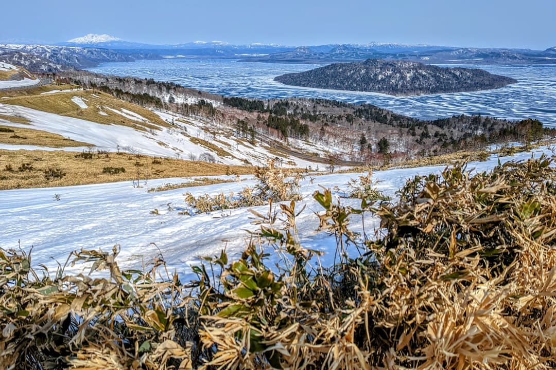 A spring thaw landscape in Hokkaido showing a frozen caldera lake with forested islands, melting snow on hillsides, and a snow-capped volcanic peak visible in the distance under clear skies. - Photo by Da-shika on Unsplash