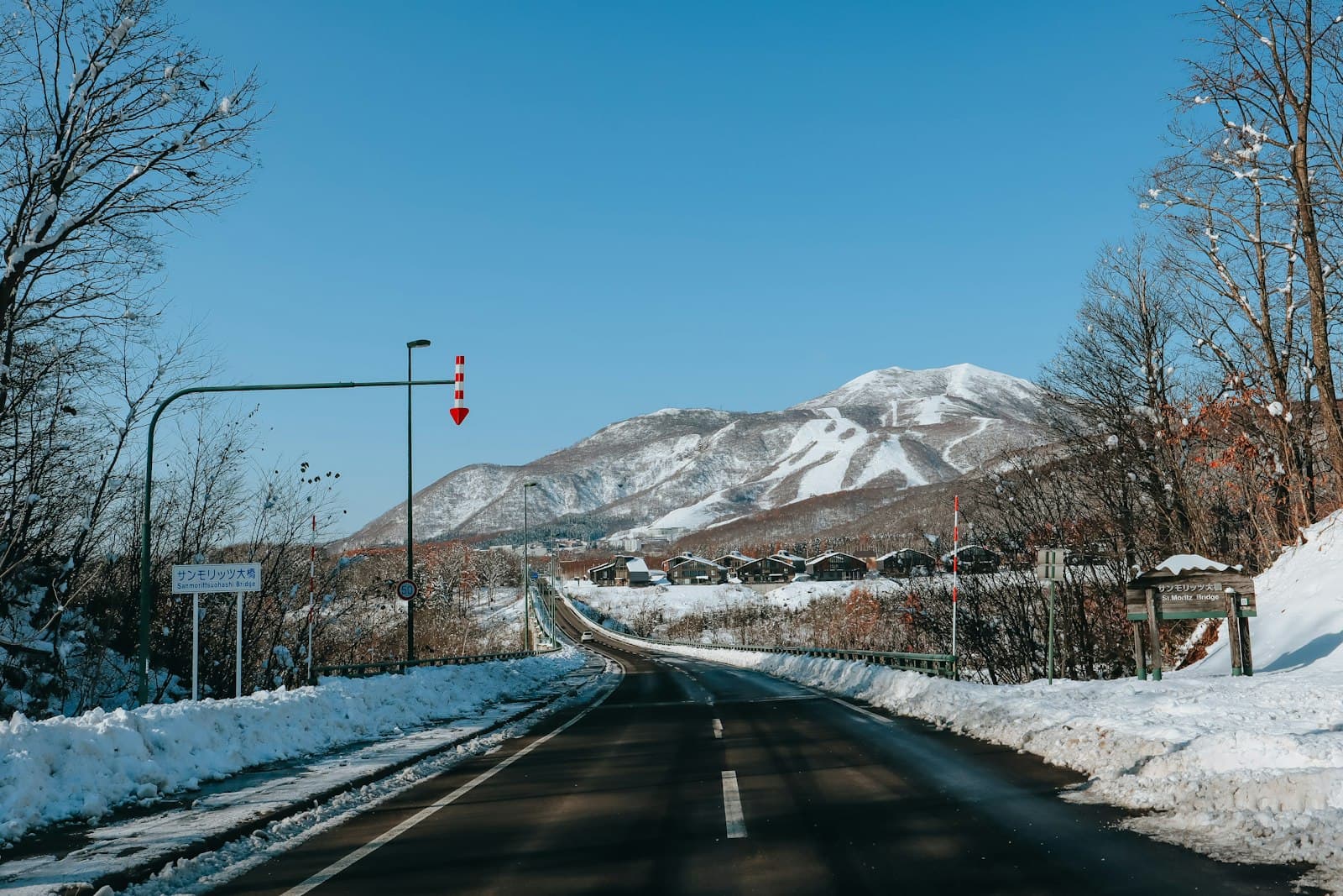 A winter mountain pass road in snow with ski resort infrastructure visible, featuring a distinctive ski slope on the mountainside in the background and Japanese resort signage. - Photo by Asmut Dante on Unsplash