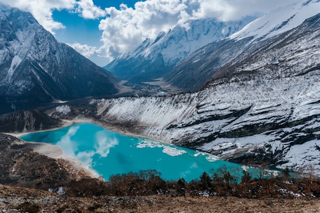 A turquoise glacial meltwater lake nestled in a high-altitude alpine valley surrounded by snow-dusted mountains and rocky terrain, with snow-capped peaks visible in the distance. - Photo by Bobby on Unsplash