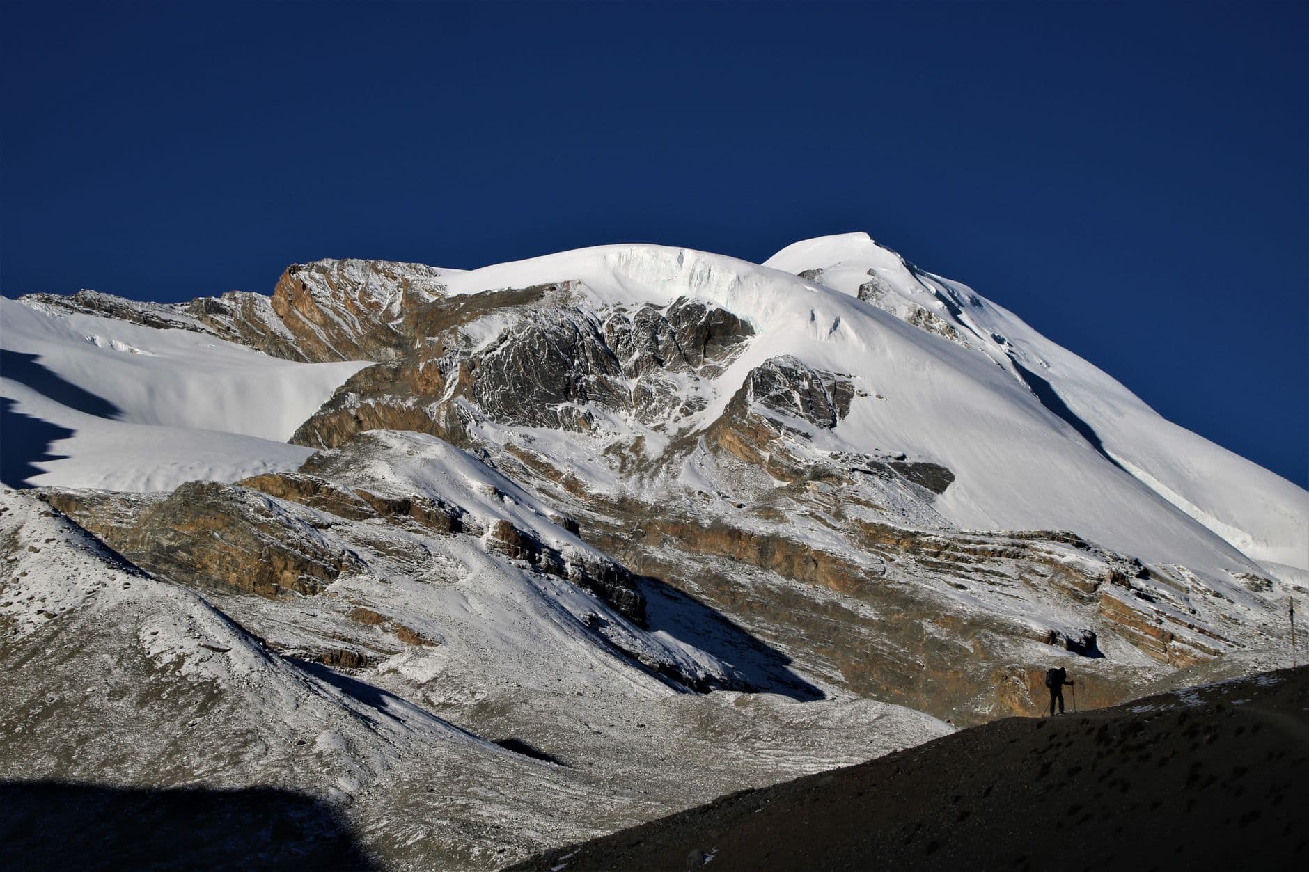 A snow-covered Himalayan peak with exposed geological strata, glaciers, and a lone trekker on the mountainside under clear blue sky. - Photo by Vertex Holiday on Pexels