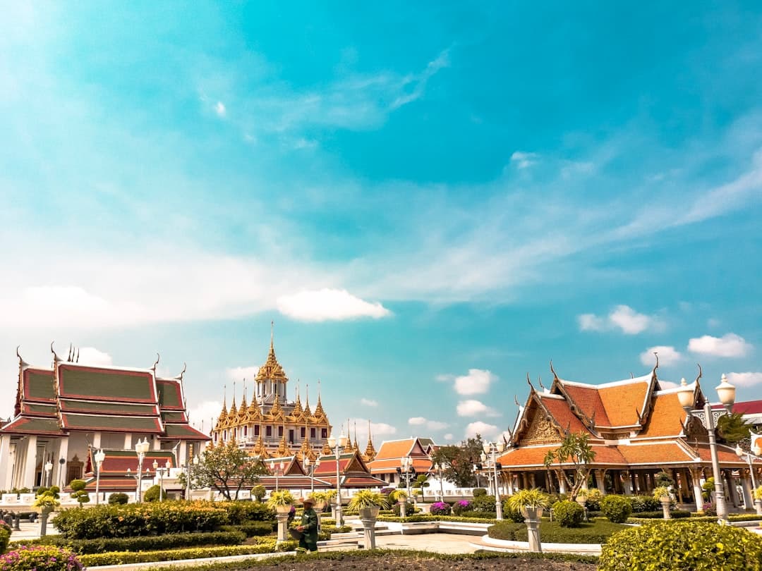A serene Thai Buddhist temple complex in Bangkok featuring multiple ornate golden and red-roofed buildings with characteristic upturned eaves, manicured gardens, and clear blue sky. - Photo by Alejandro Cartagena 🇲🇽🏳🌈 on Unsplash