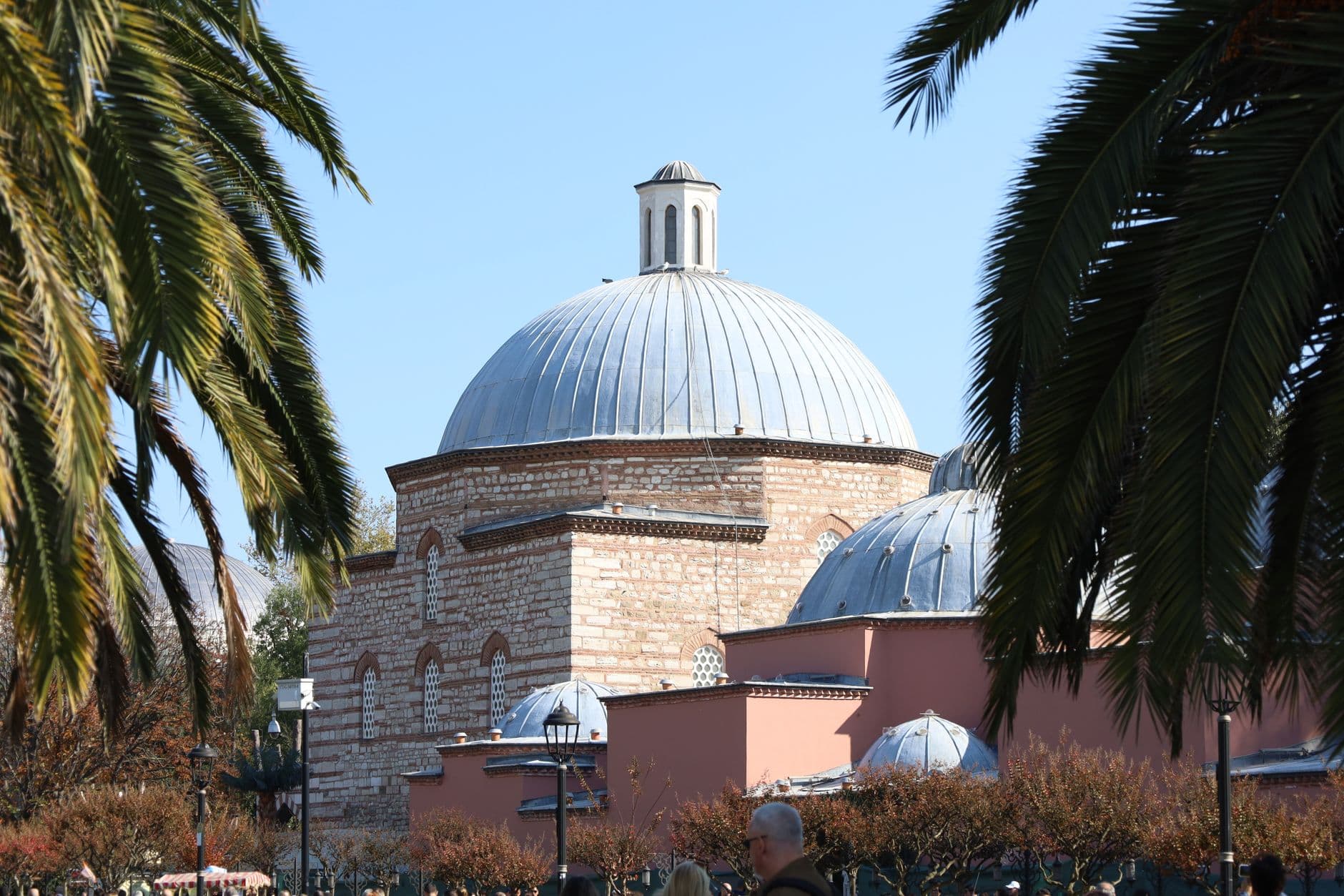 A Ottoman-style domed building with red brick walls, lead domes, and lantern towers, framed by palm fronds, depicting a historic Turkish monument in an urban setting. - Photo by Serg Alesenko on Pexels