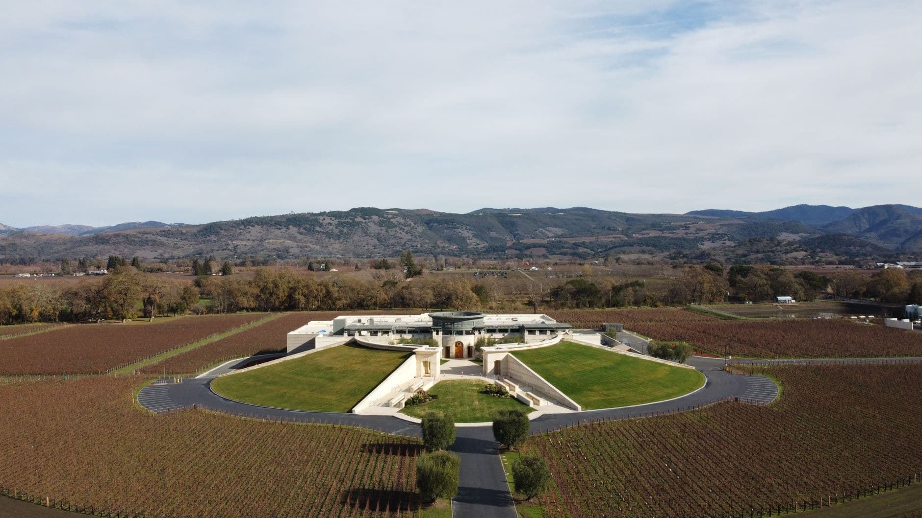 A modern, architecturally distinctive winery building with curved green roofing built into a hillside, surrounded by vineyard rows, photographed from aerial perspective with Napa Valley hills visible in the background. - Photo by Riccardo Zerbinati on Pexels