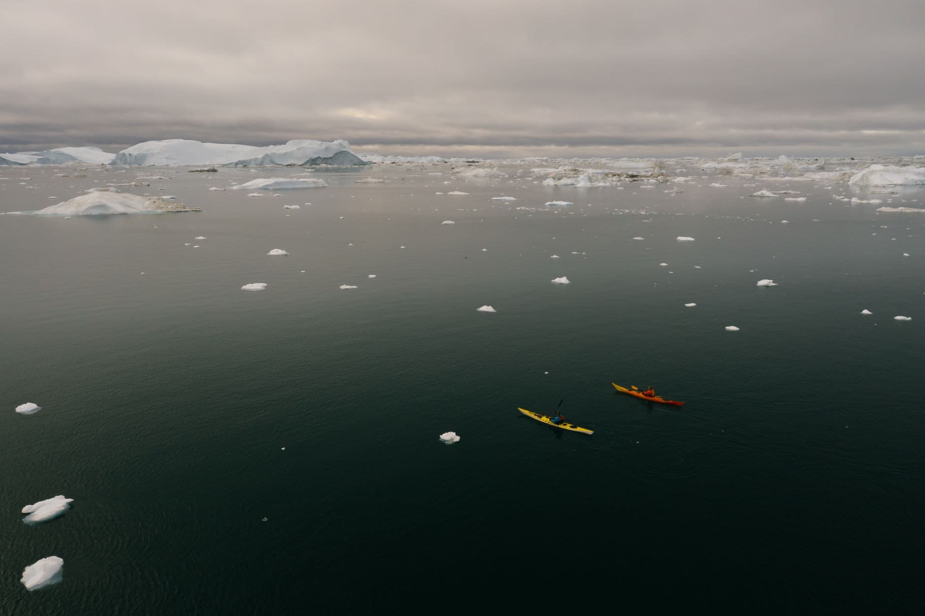 Two sea kayakers paddle through scattered icebergs and calved ice fragments in a cove surrounded by glaciated mountains, with larger ice shelves visible on the horizon. - Photo by Mikhail Nilov on Pexels