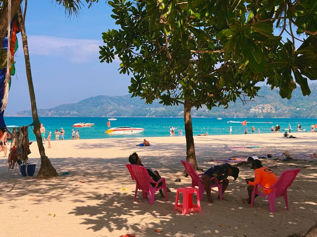 A busy tropical beach in Phuket with tourists swimming and relaxing under colorful plastic chairs beneath mangrove trees, with speedboats anchored in the turquoise water and forested hills rising behind the bay. - Photo by Max Bvp on Unsplash