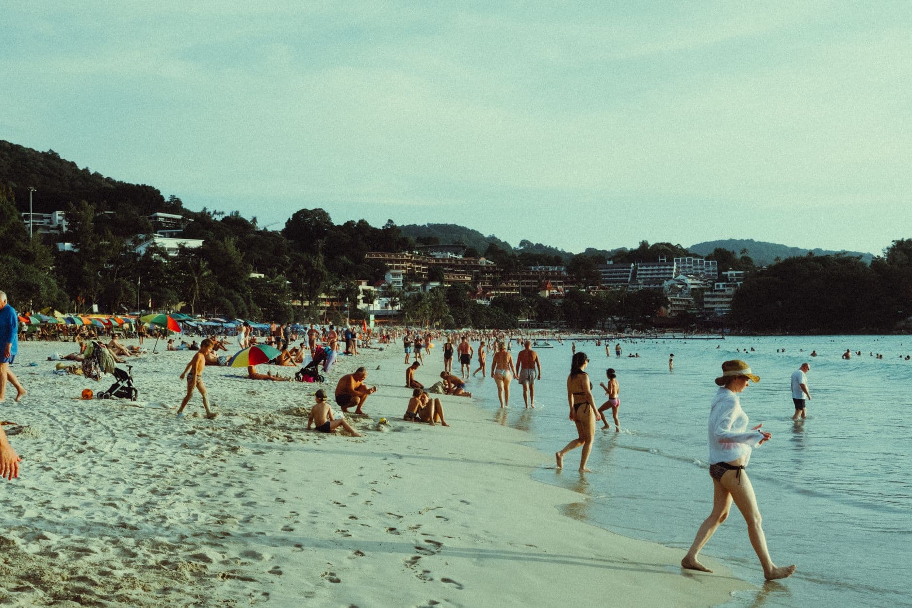 A crowded tropical beach scene with hundreds of swimmers and beachgoers enjoying shallow turquoise water, with multi-story resort hotels and residential buildings built into forested hills behind the beach. - Photo by Gizem Çelebi on Pexels