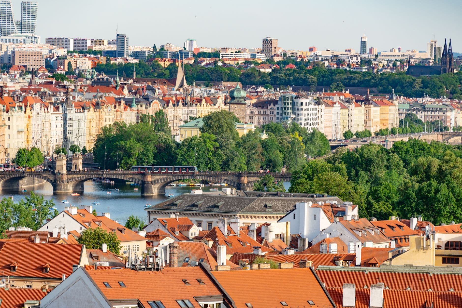 A panoramic cityscape of Prague taken from an elevated vantage point showing the Vltava River with Charles Bridge, colorful old-town buildings with terracotta roofs, and Prague Castle and spires dominating the skyline beyond. - Photo by Diego F. Parra on Pexels
