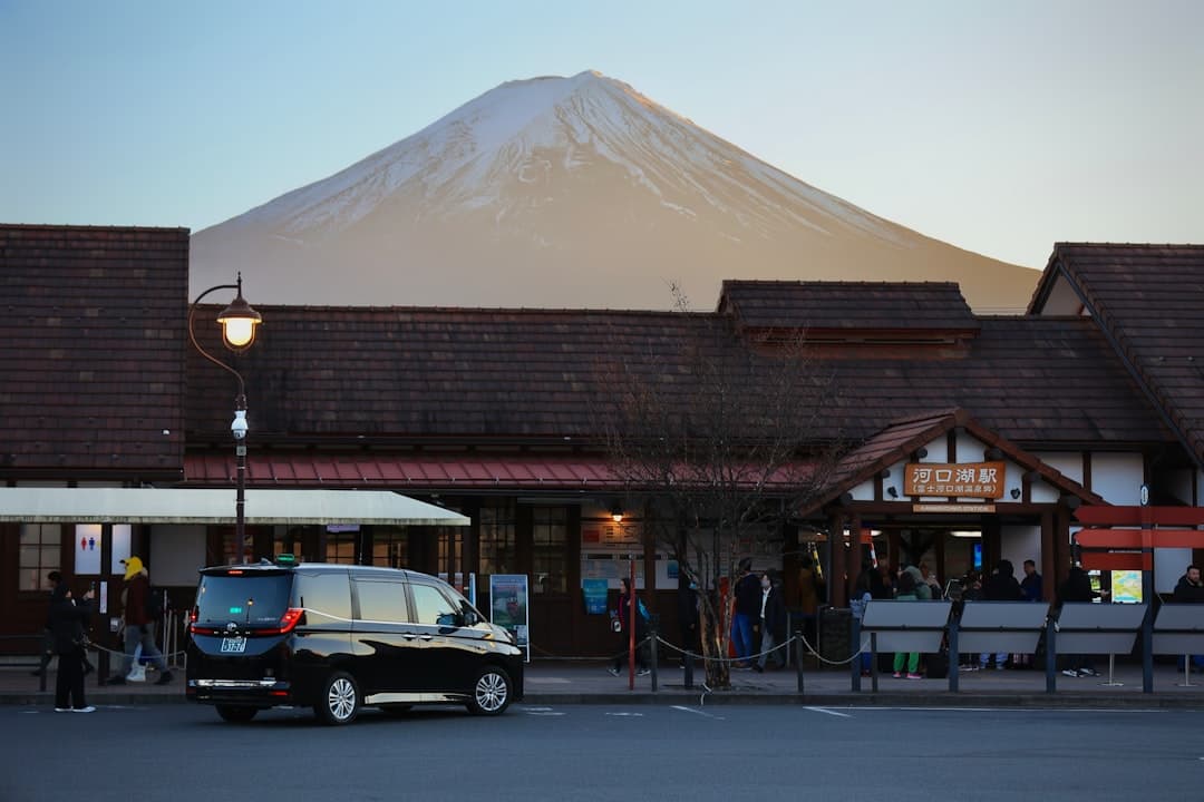 A train station area at Lake Kawaguchi with Mount Fuji snow-capped peak visible in the background, featuring traditional Japanese architecture, parked vehicles, and pedestrians. - Photo by Jiachen Lin on Unsplash