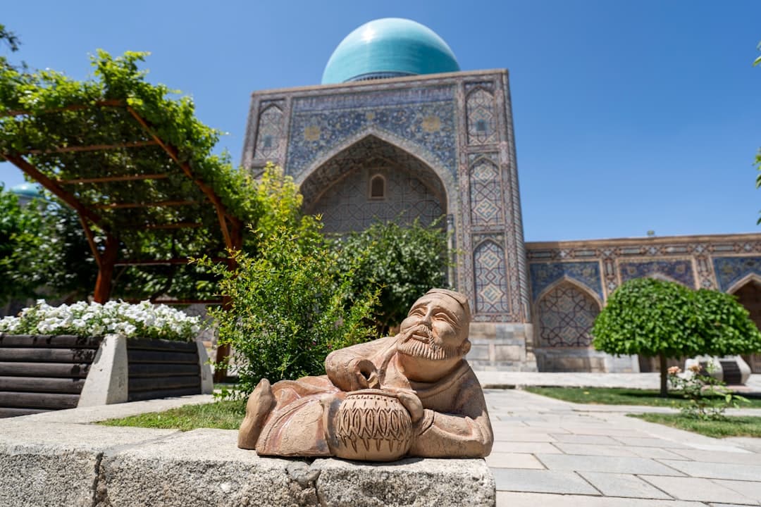 A terracotta sculptural figure in a courtyard of what appears to be the Registan in Samarkand, with a historic medrese building featuring a turquoise dome and intricate tilework visible in the background. - Photo by Hendrik Morkel on Unsplash