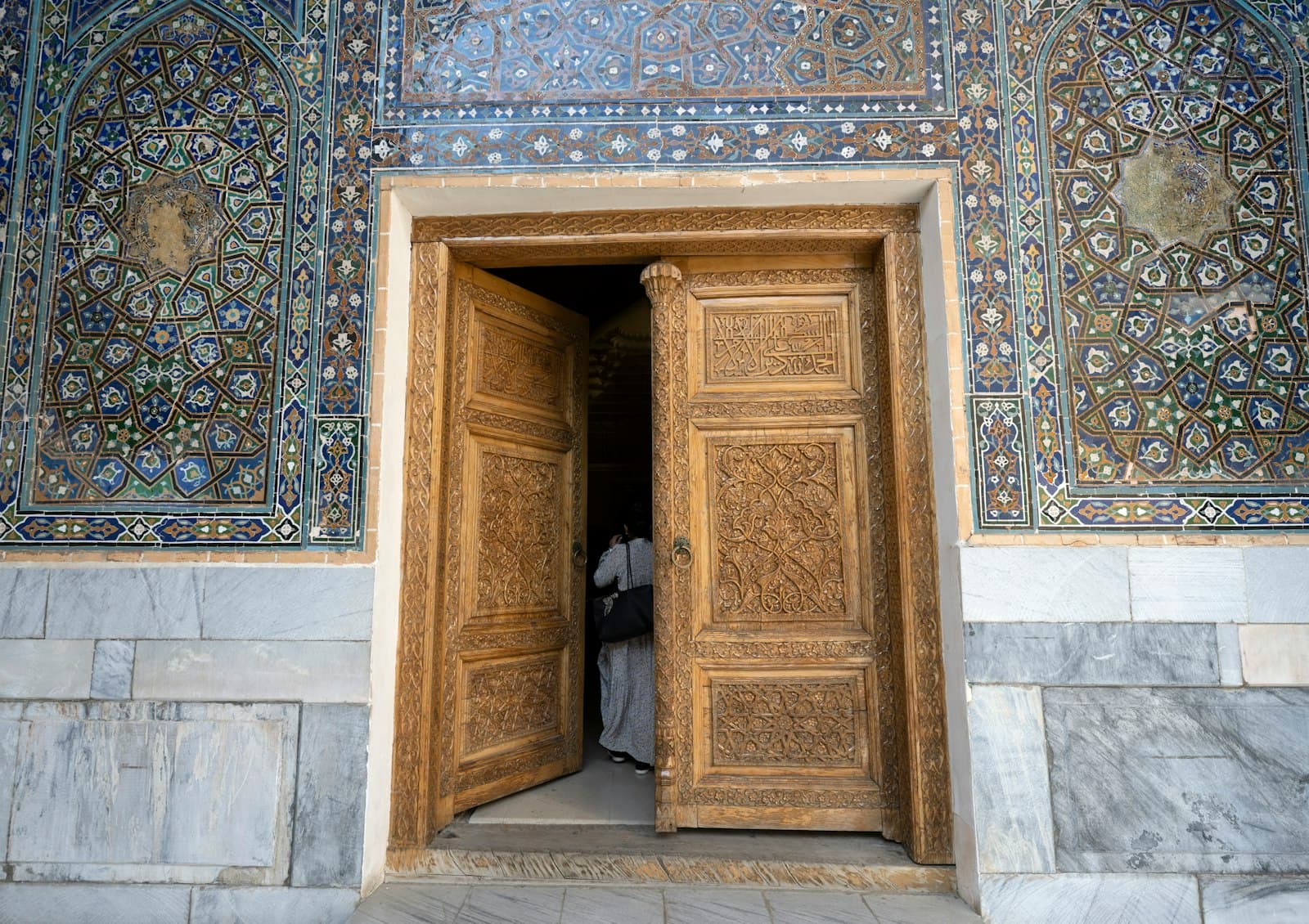 An intricately decorated doorway with ornate carved wooden doors opens into a darkened interior, framed by spectacular tilework featuring Islamic geometric patterns in blue, white, and gold on the surrounding walls. - Photo by Dave Meckler on Unsplash