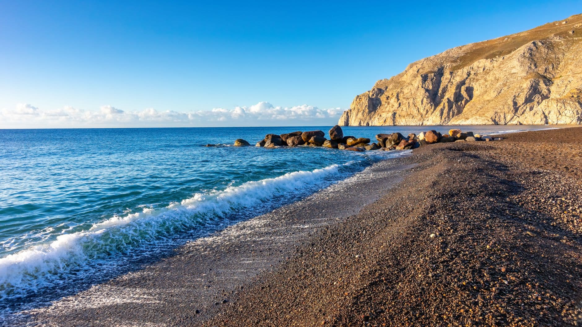 A scenic black sand beach with calm blue waters, white-capped waves, dark volcanic rocks, and towering golden-brown volcanic cliffs under clear sky. - Photo by Wojciech Wyszkowski on Pexels
