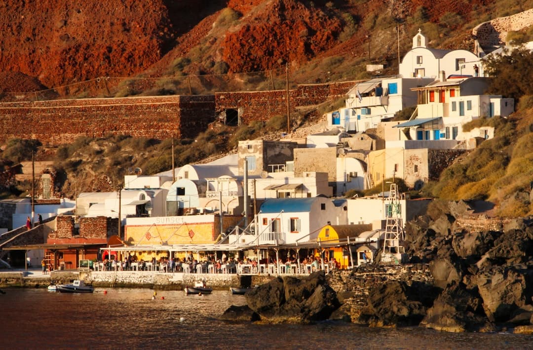A waterfront village built into dramatic red-brown volcanic cliffs, featuring whitewashed buildings with blue accents, a colorful waterfront taverna, and traditional Cycladic architecture typical of the Aegean islands. - Photo by Marc Snailum on Unsplash