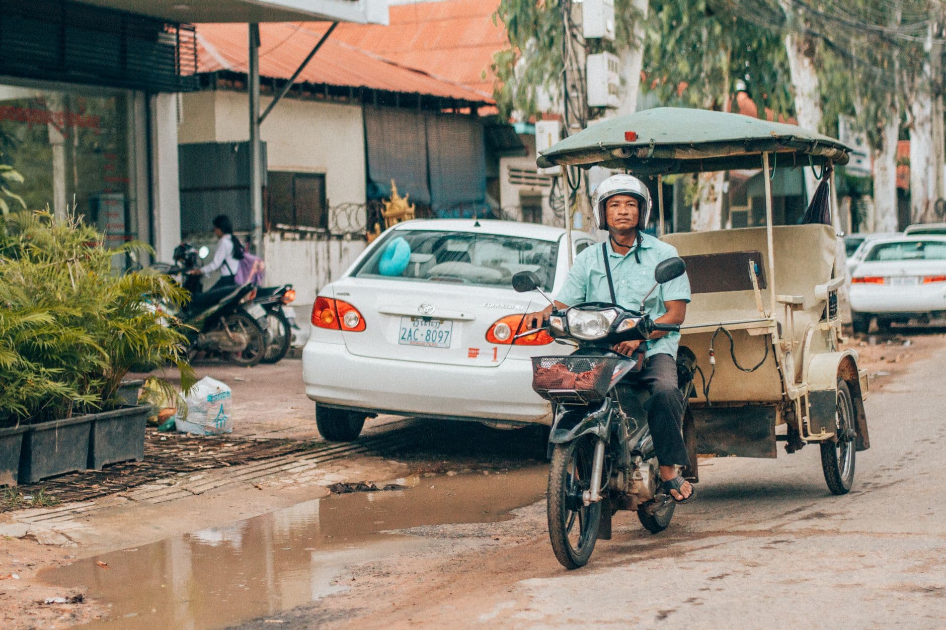 A man on a motorized trike/tuk-tuk towing a cargo trailer navigates a muddy street past parked vehicles and simple buildings in what appears to be a Cambodian provincial town. - Photo by Humphrey Muleba on Pexels