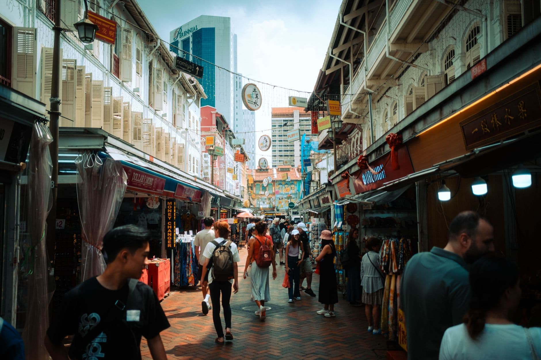 A bustling street market or shopping area in Singapore's Chinatown, lined with traditional shophouses featuring Chinese signage, red lanterns, and colorful storefronts, with pedestrians browsing goods and modern buildings visible in the background. - Photo by Fabian Reck on Pexels
