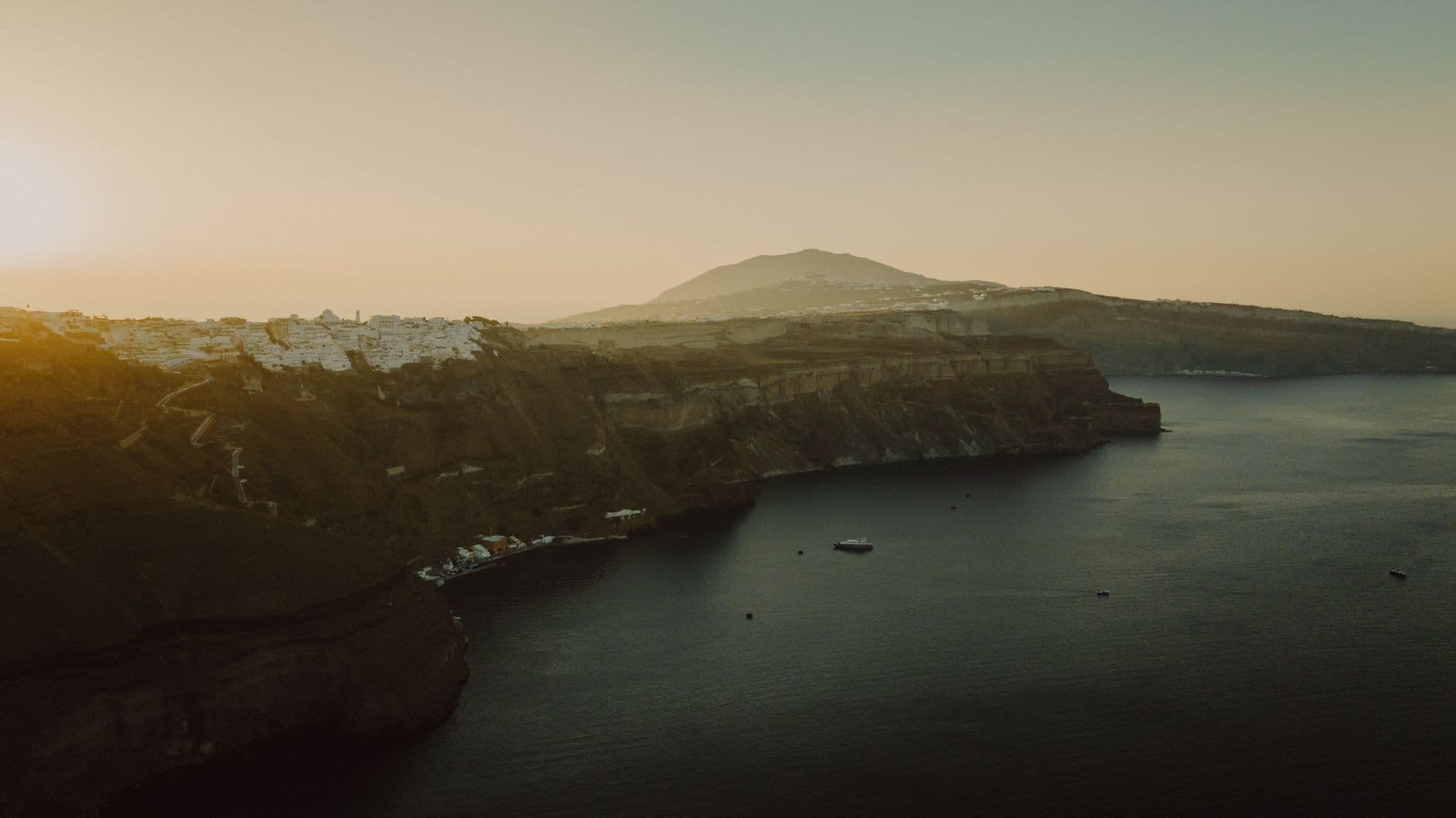 A sunset or golden-hour view of Santorini's caldera showing dramatic dark volcanic cliffs, whitewashed buildings on the cliff rim, anchored boats in the bay, and a volcanic mountain rising in the distance across the water. - Photo by Ushindi Namegabe on Pexels