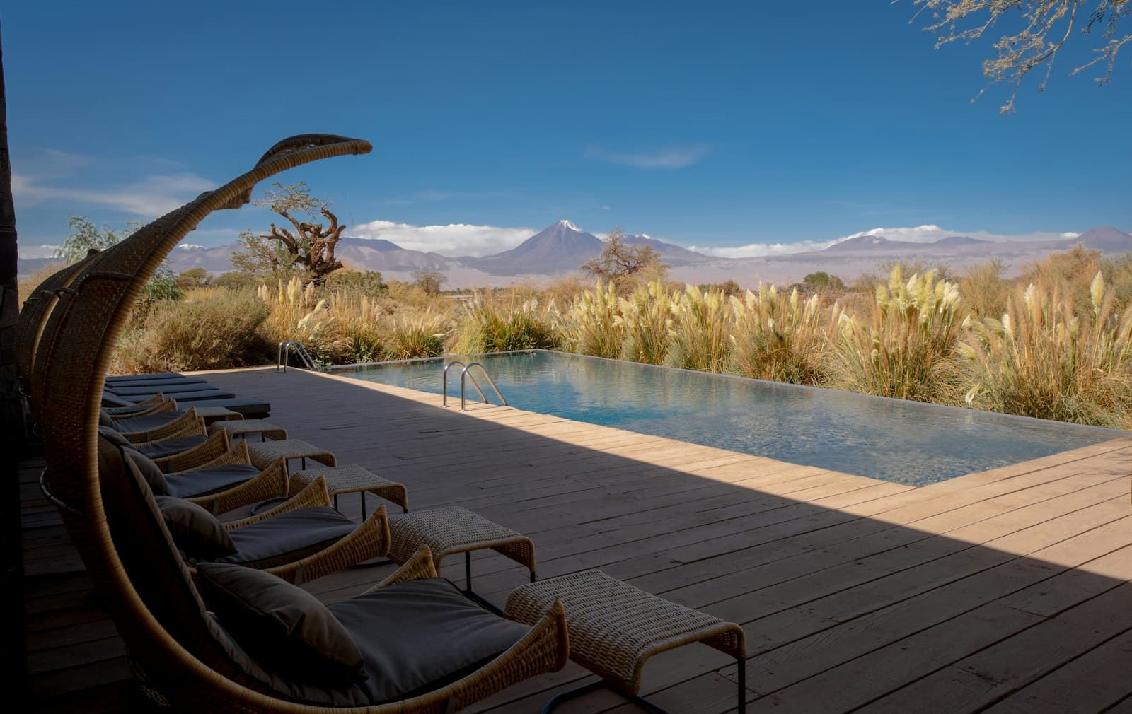 A luxury poolside deck with woven lounge chairs overlooking an infinity pool, with the arid Atacama Desert and distant snow-capped volcanoes visible beyond sparse vegetation. - Photo by Diego Marín on Unsplash