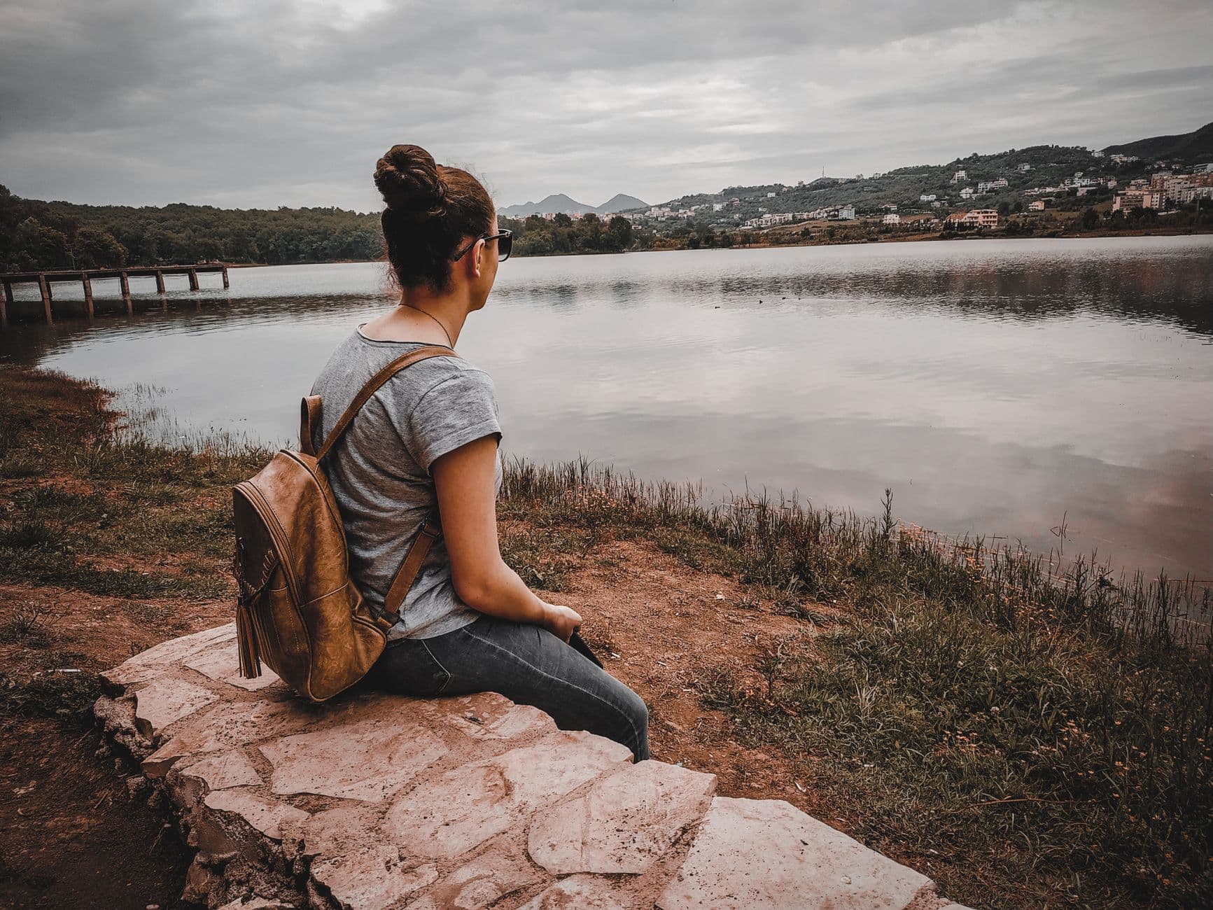 A solo traveler sitting on rocks overlooking a calm lake or reservoir with forested hillsides and residential development visible across the water. - Photo by Besjan Bardhollari on Pexels