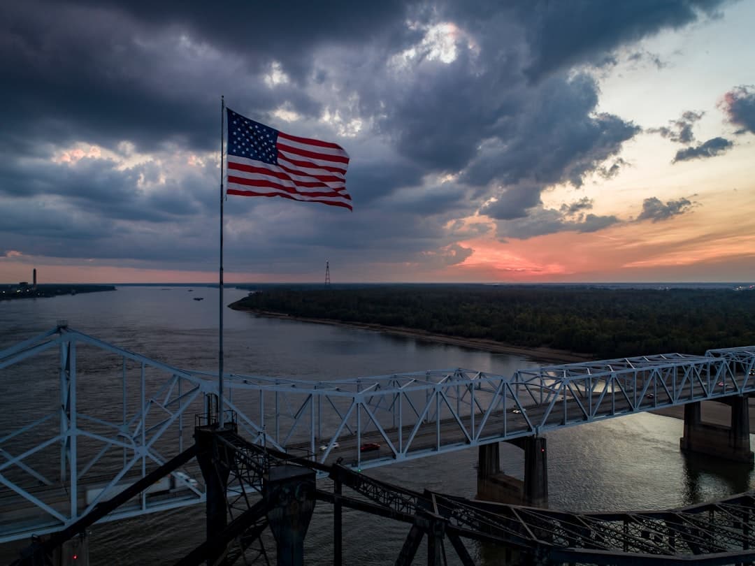An American flag flies at sunset or sunrise above the Lower Mississippi River, with a white metal railing and observation platform in the foreground overlooking a wide bend in the river with forested banks. - Photo by Justin Wilkens on Unsplash