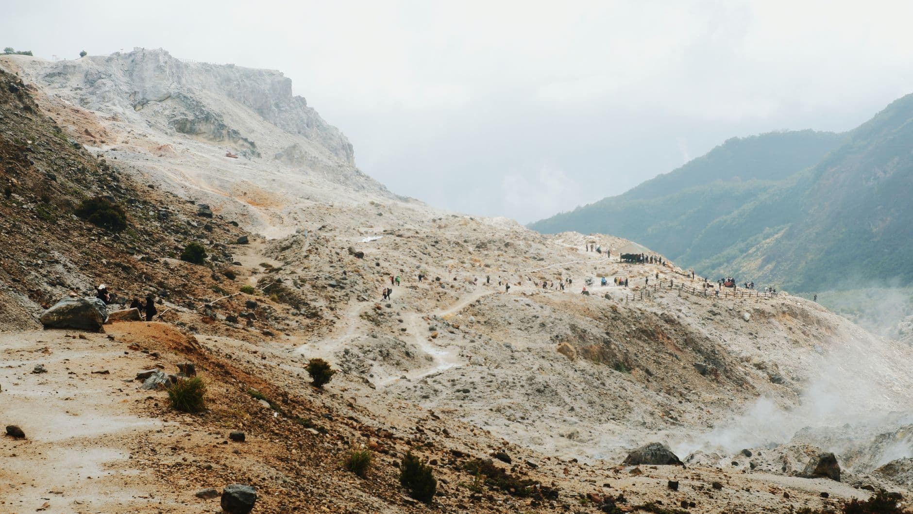 Trekkers navigate a barren, mineral-rich volcanic crater landscape with steep ash and sulfur deposits, safety railings visible on the ridge above, and a valley with green vegetation visible in the distance. - Photo by Nurul Sakinah Ridwan on Pexels
