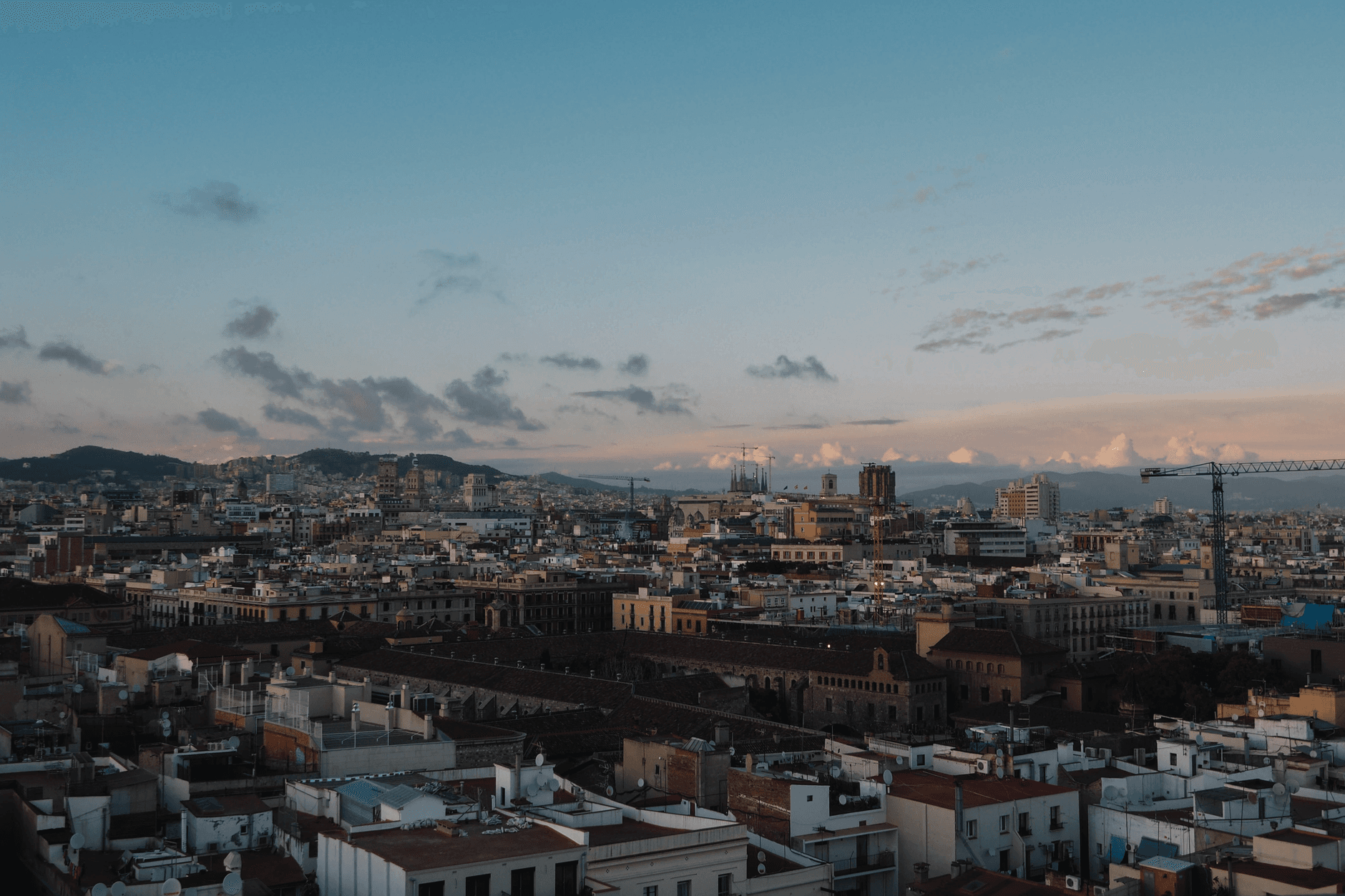 A sunset cityscape photograph taken from a high vantage point showing Barcelona's dense urban sprawl with a mix of historic and modern architecture, hills in the distance, and dramatic clouds lit by golden hour light. - Photo by Lia L. on Pexels