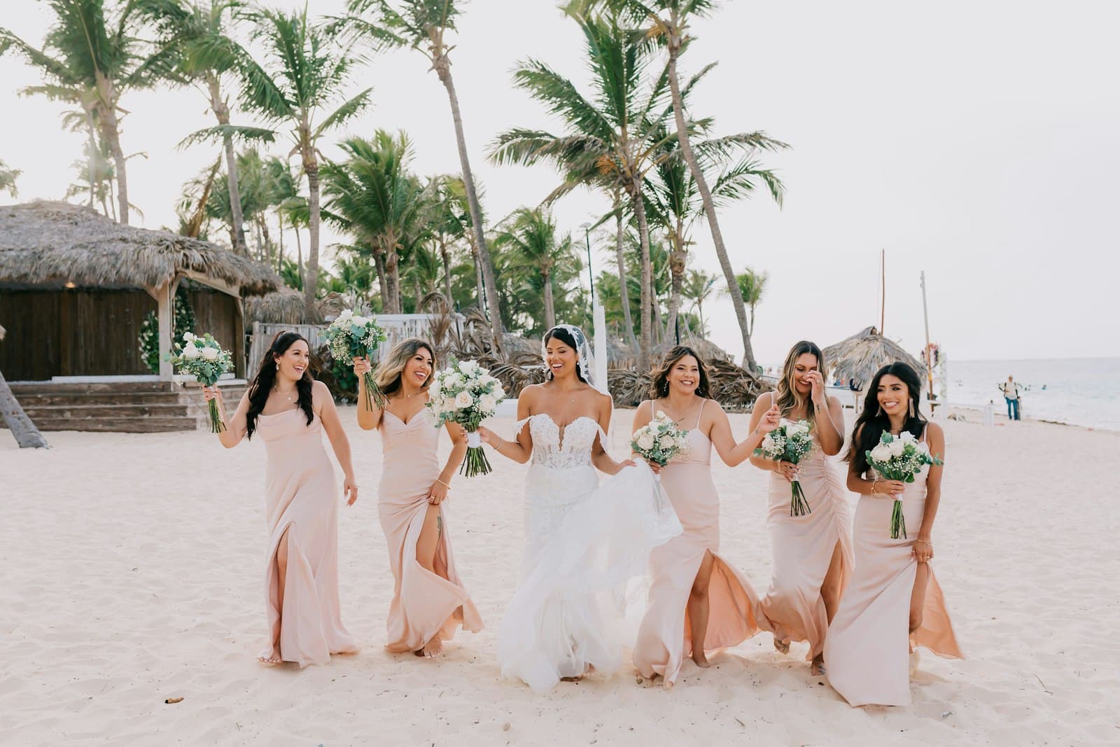 A bride and bridesmaids walk together on a white sand beach in front of thatched-roof beach structures and palm trees, holding white and green bouquets during what appears to be a wedding celebration. - Photo by Mikaela San Juan on Unsplash