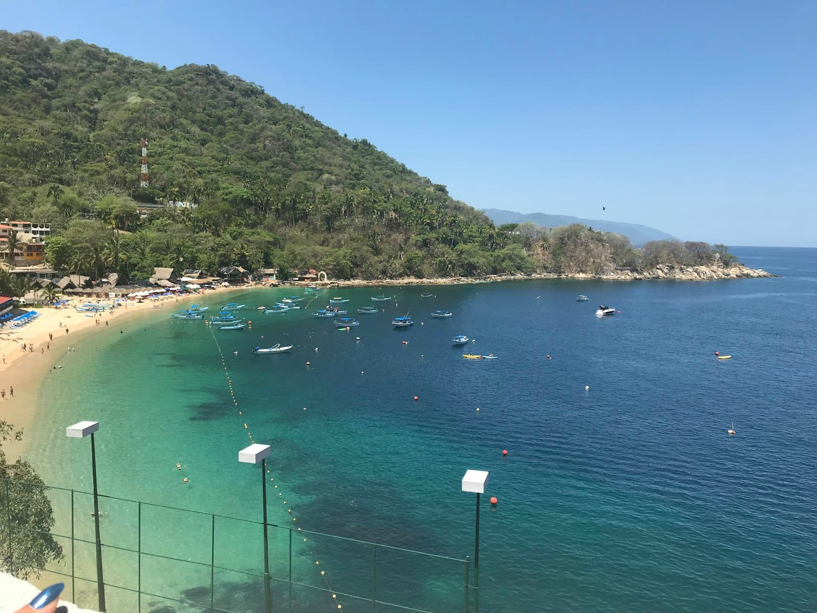 A scenic cove with turquoise shallow waters, a sandy beach lined with palapa umbrellas and loungers, dense tropical vegetation covering a steep hillside, and numerous small fishing boats moored in the bay. - Photo by Alondra López on Unsplash