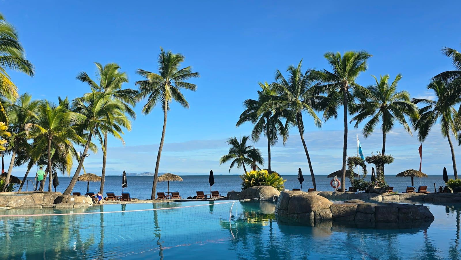 A luxury beachfront resort pool with palm trees, large boulders, and ocean view overlooking Denarau Island's coastal waters. - Photo by Louise Smith on Unsplash
