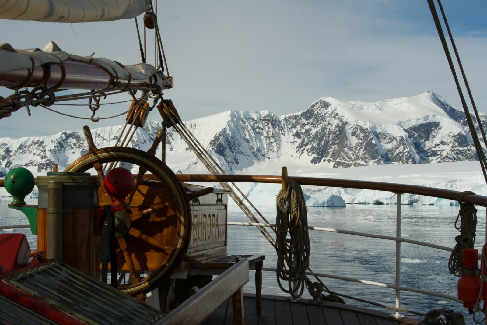 View from the deck of an expedition cruise ship (Europa) showing the ship's wheel and rigging with snow-covered mountains and icebergs of the Gerlache Strait visible across the water. - Photo by Torsten Dederichs on Unsplash