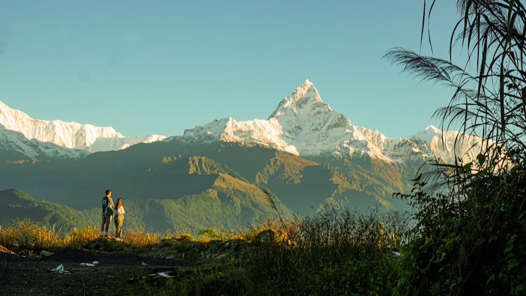 Two hikers stand on a hillside viewing a snow-capped mountain range dominated by a distinctive pyramidal peak, with forested valleys in the foreground and misty lower elevations. - Photo by Anja Lee Ming Becker on Unsplash