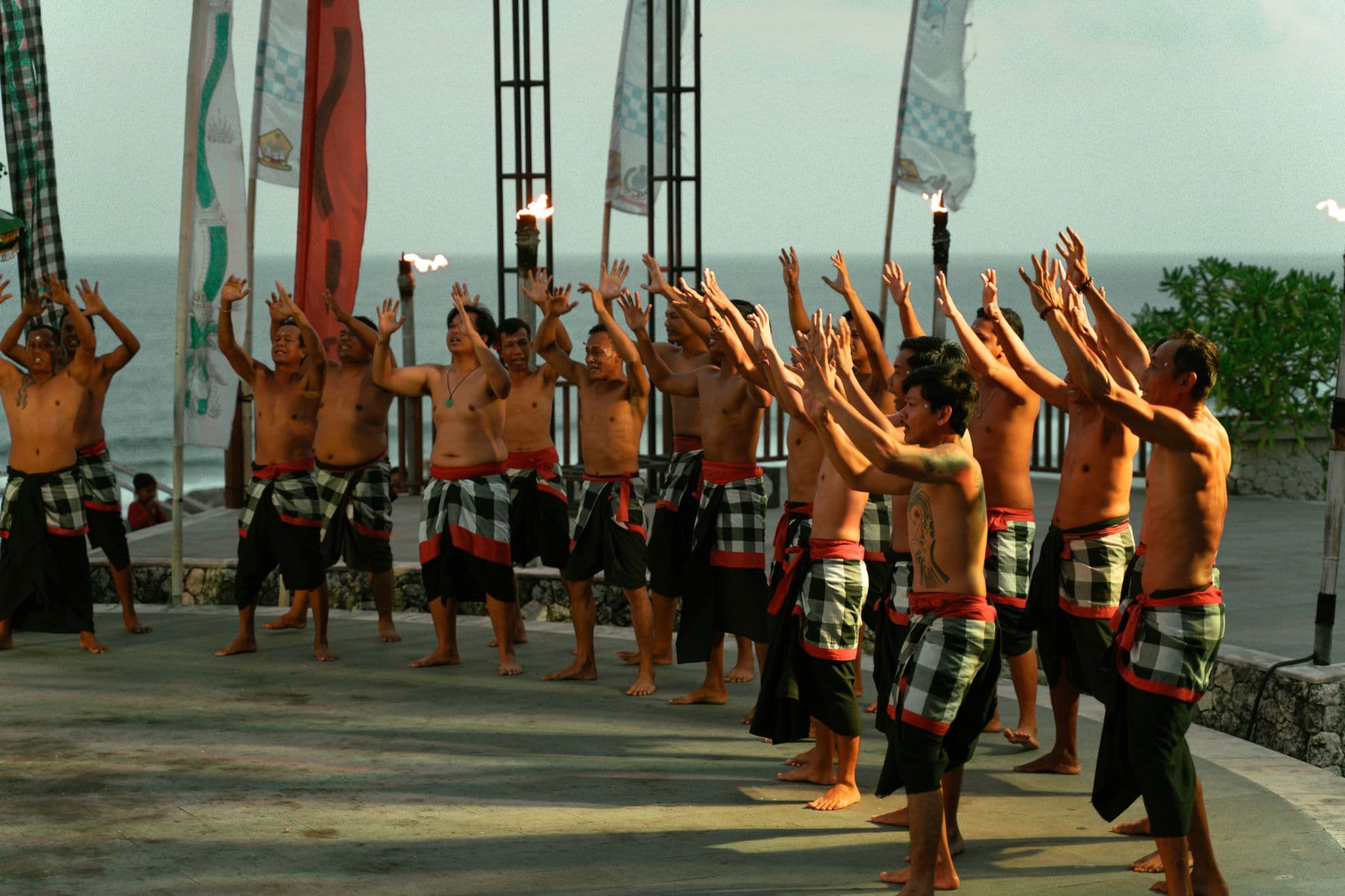 A group of shirtless young Balinese men in traditional sarongs perform a synchronized hand gesture or ceremonial movement on a coastal platform overlooking the ocean, with decorative flags and torches visible in the background. - Photo by Ilham Zovanka on Pexels
