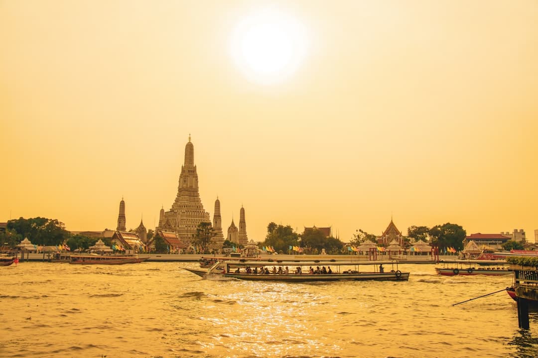A golden-hour photograph of Wat Arun temple across the Chao Phraya River in Bangkok, with traditional longtail boats and riverside structures visible. - Photo by Steven Wilcox on Unsplash