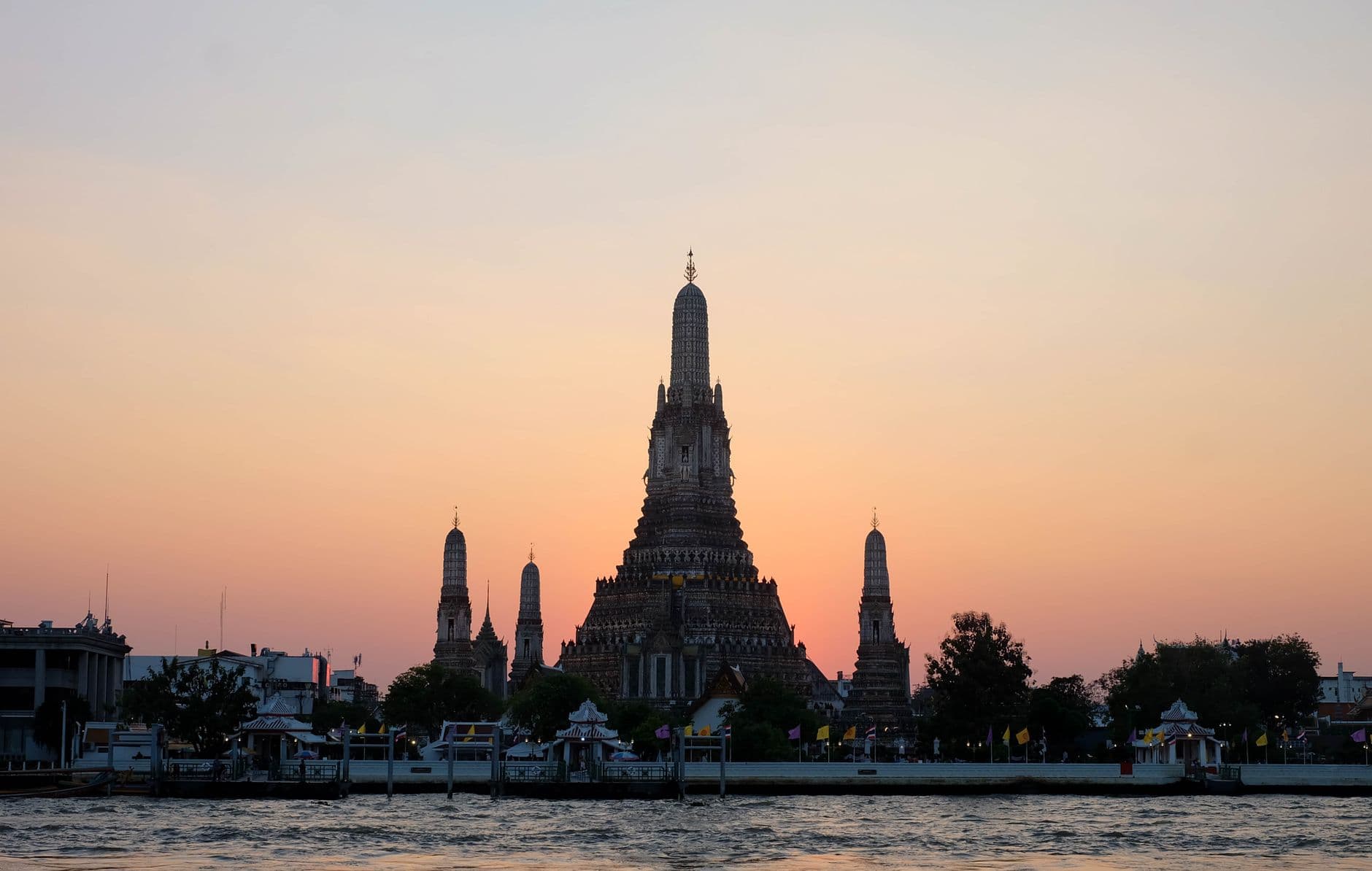 Sunset view of Wat Arun and the Bangkok waterfront along the Chao Phraya River, with the temple's central spire silhouetted against a warm orange and pink sky. - Photo by Margarita K on Pexels