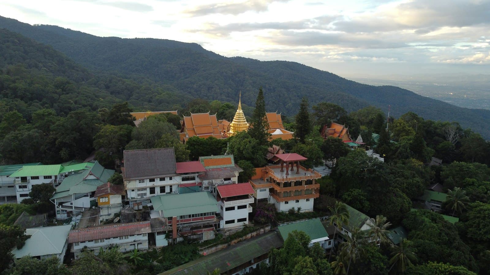 An aerial view of a Buddhist temple complex nestled in forested mountains, surrounded by modern Thai buildings with metal roofing, showing the characteristic architecture and landscape of northern Thailand. - Photo by Mike Holp on Unsplash