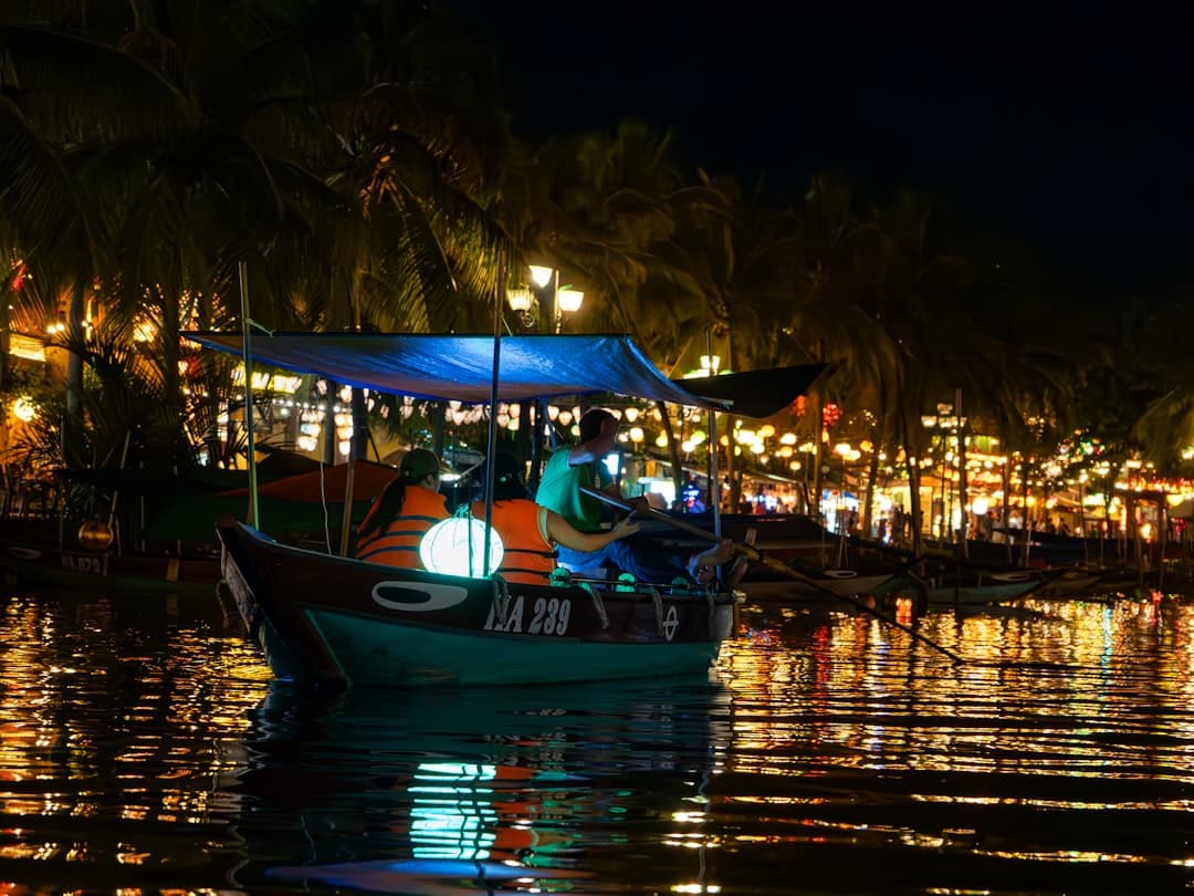 A traditional wooden fishing boat lit at night on the Thu Bon River in Hoi An, with palm trees and the illuminated waterfront promenade of the Ancient Town visible in the background. - Photo by Juup Schram on Unsplash