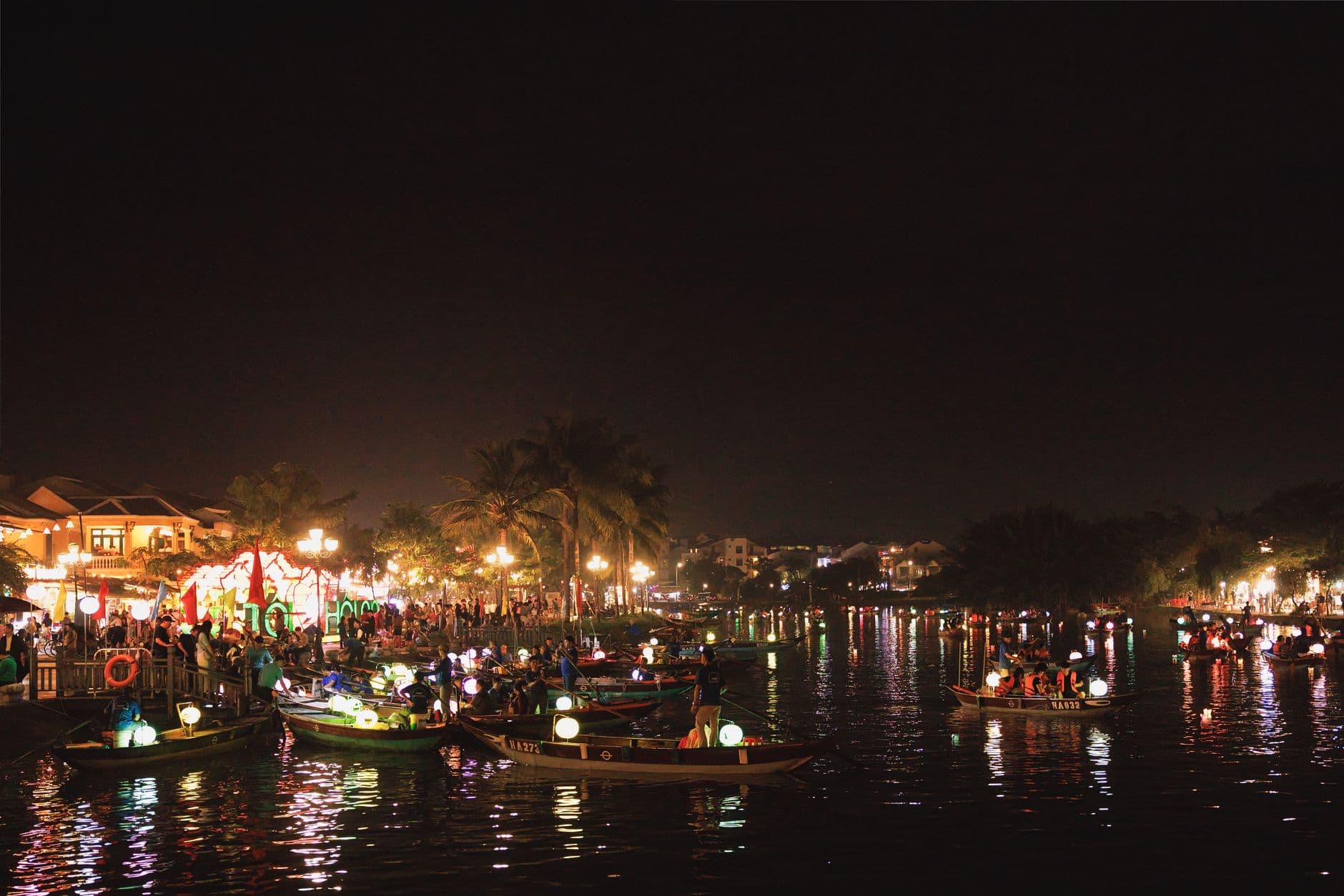 Night scene of illuminated traditional Vietnamese fishing boats on a river with lanterns, waterfront buildings, palm trees, and crowds of visitors along the shore during what appears to be a festival or evening market event. - Photo by Fernando B M on Pexels