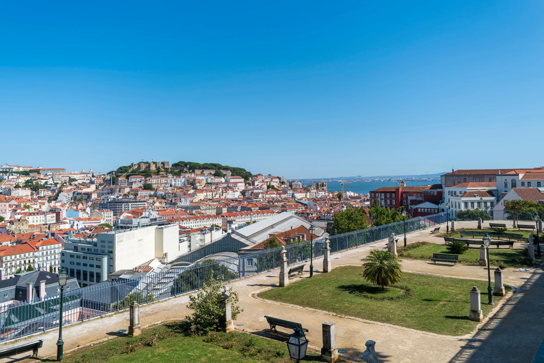 A viewpoint overlooking Lisbon's cityscape with red-tiled roofs, São Jorge Castle on the hilltop, the Tagus River in the distance, and a modern viewing platform with benches in the foreground. - Photo by Bob Jenkin on Pexels